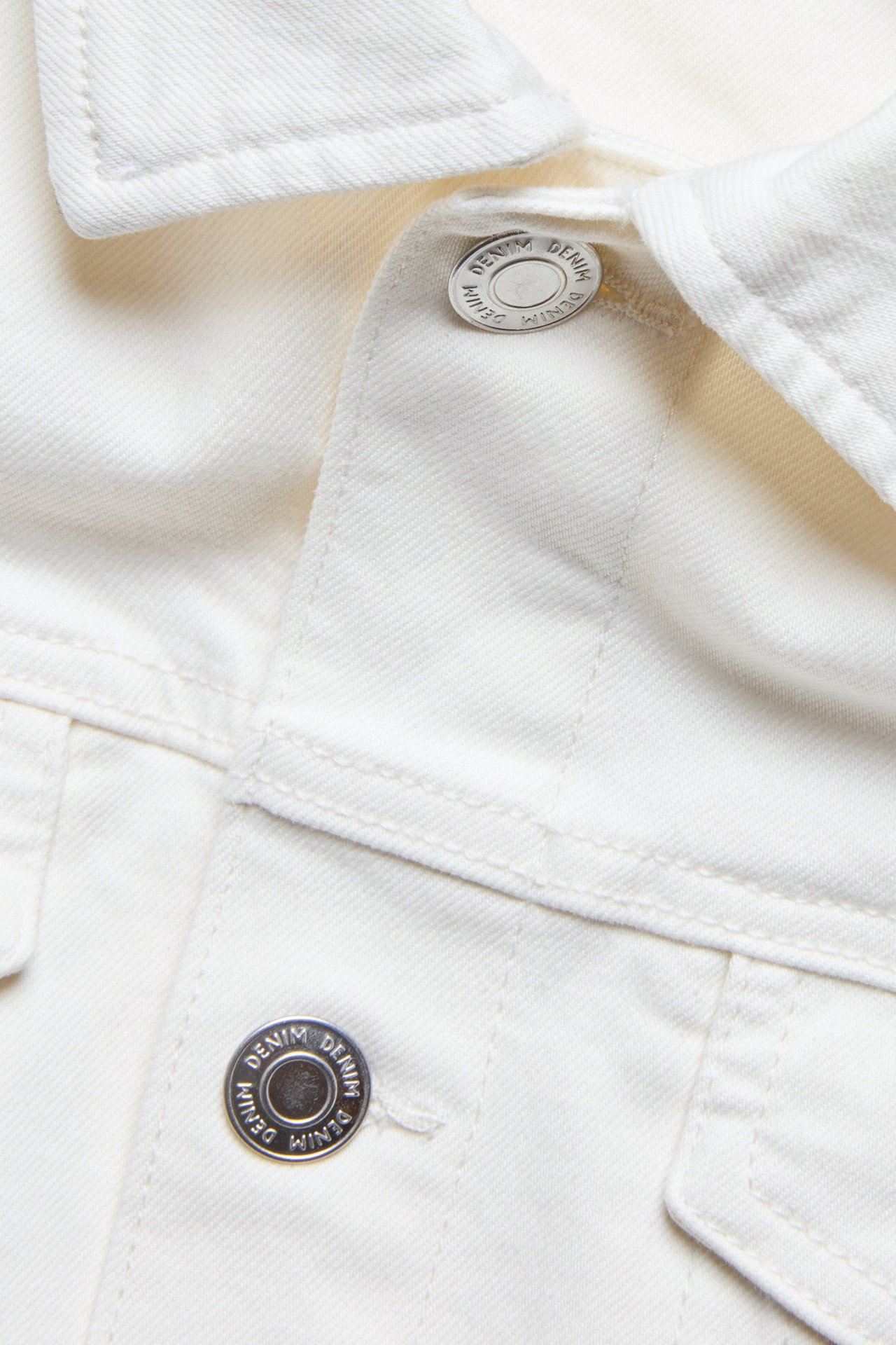 Zbliżenie beżowej kurtki twillowej, ukazujące srebrne guziki 'DENIM' z przodu.
