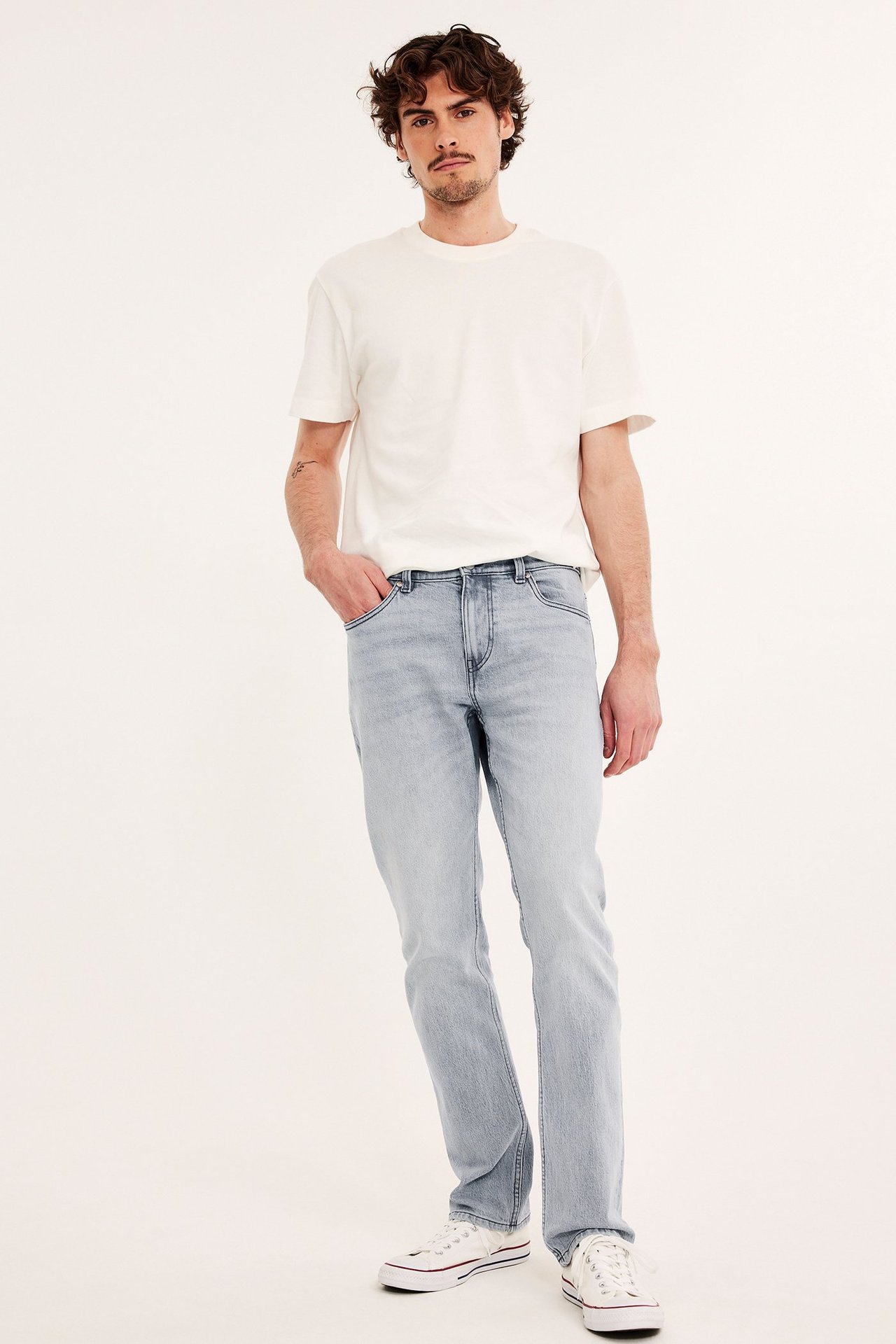 Widok z przodu mężczyzny w jasnoszarych jeansach regular fit, kremowym t-shircie i białych trampkach.
