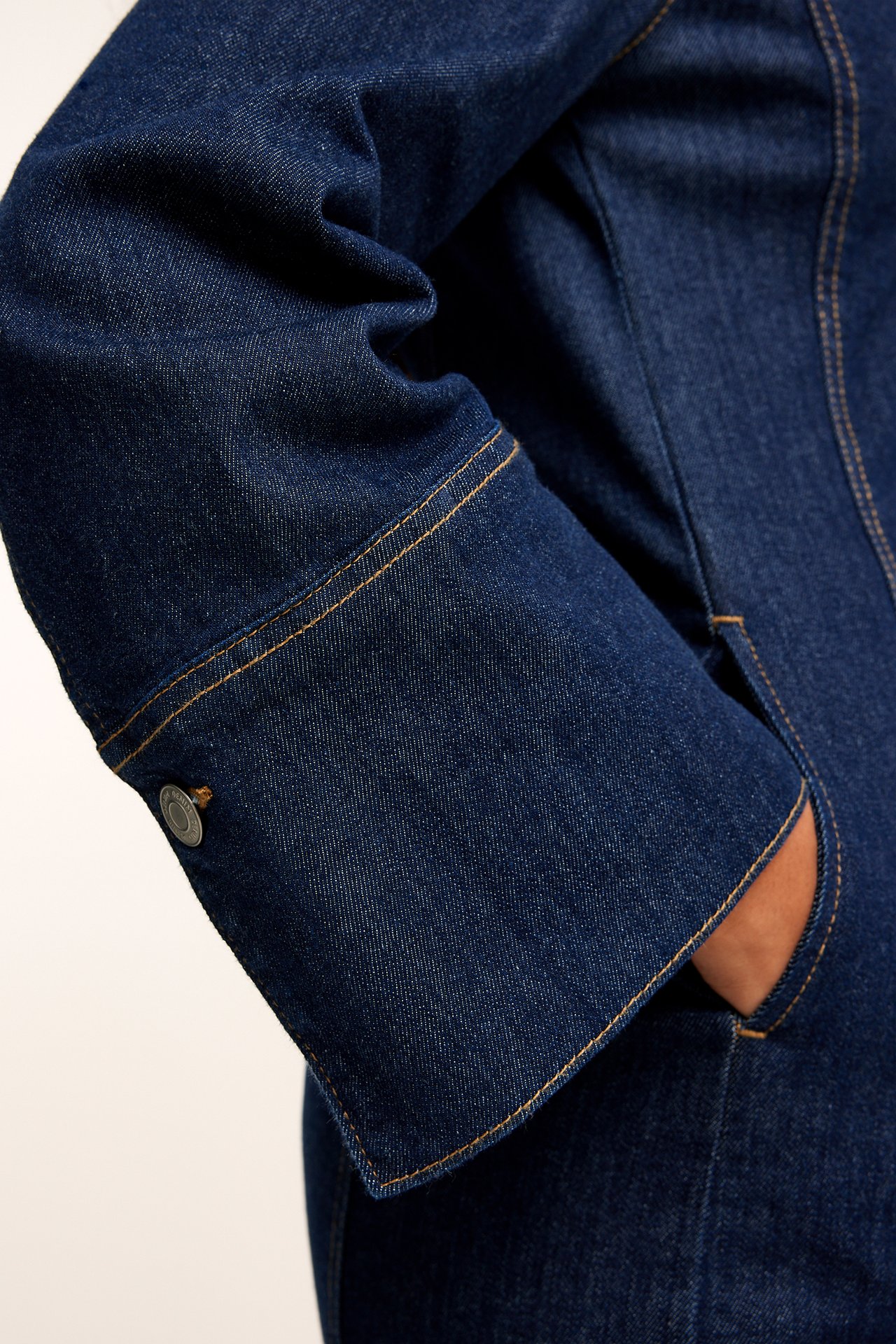 Lähikuva naisen tummasta denim-mekosta, jossa hiha, oranssit tikkaukset, hopeanappi ja käsi taskussa.