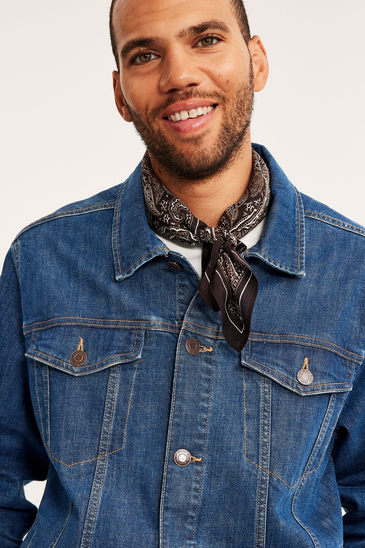 Närbild framifrån på en man i blå jeansjacka, vit t-shirt och svart paisleymönstrad bandana.