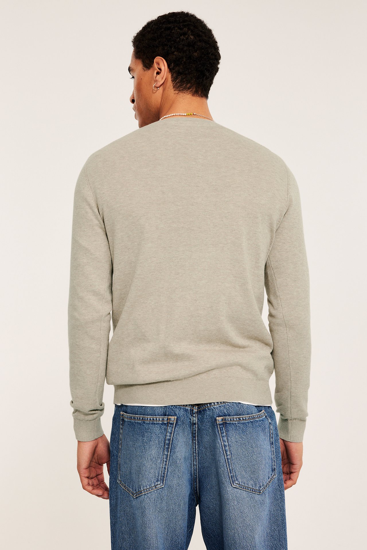 Mann, bakfra, i en beige melert finstrikket bomullsgenser, blå jeans og et perlekjede.
