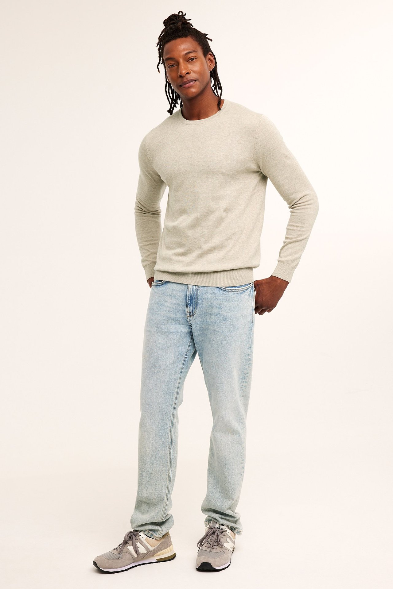 Mies edestäpäin yllään beigenvärinen meleerattu hienoneuleinen puuvillaneule, vaaleansiniset farkut ja harmaat lenkkarit.