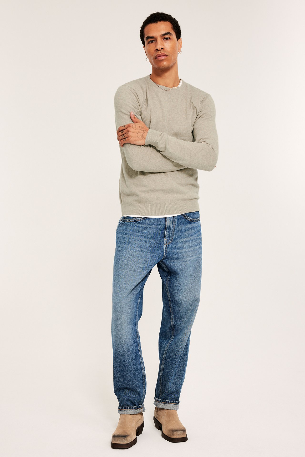Mann forfra i en beigemelert finstrikket bomullsgenser, blå jeans og beige boots.