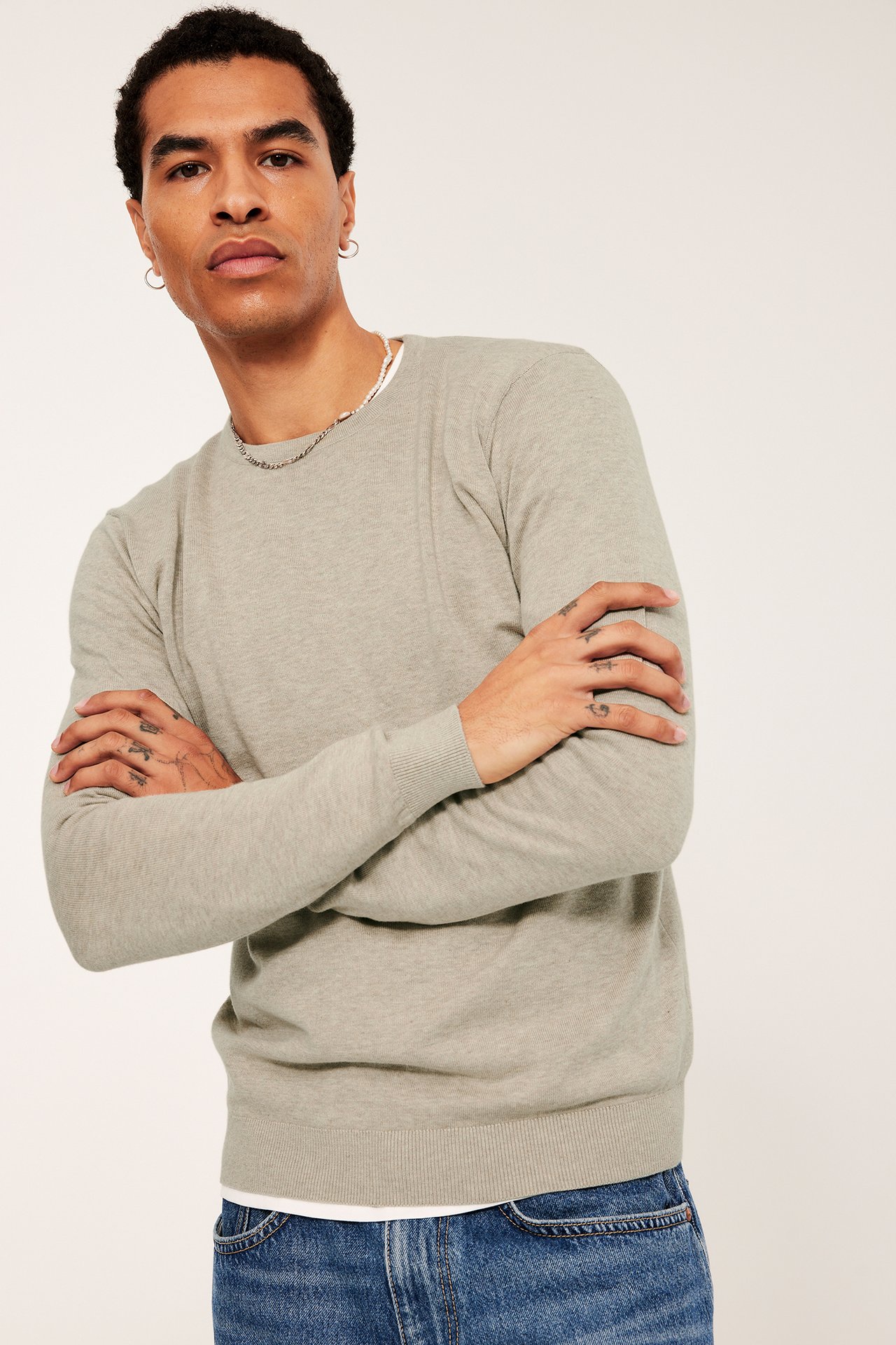 Framsida: Man i beige melerad finstickad bomullströja, vit t-shirt och blå jeans.