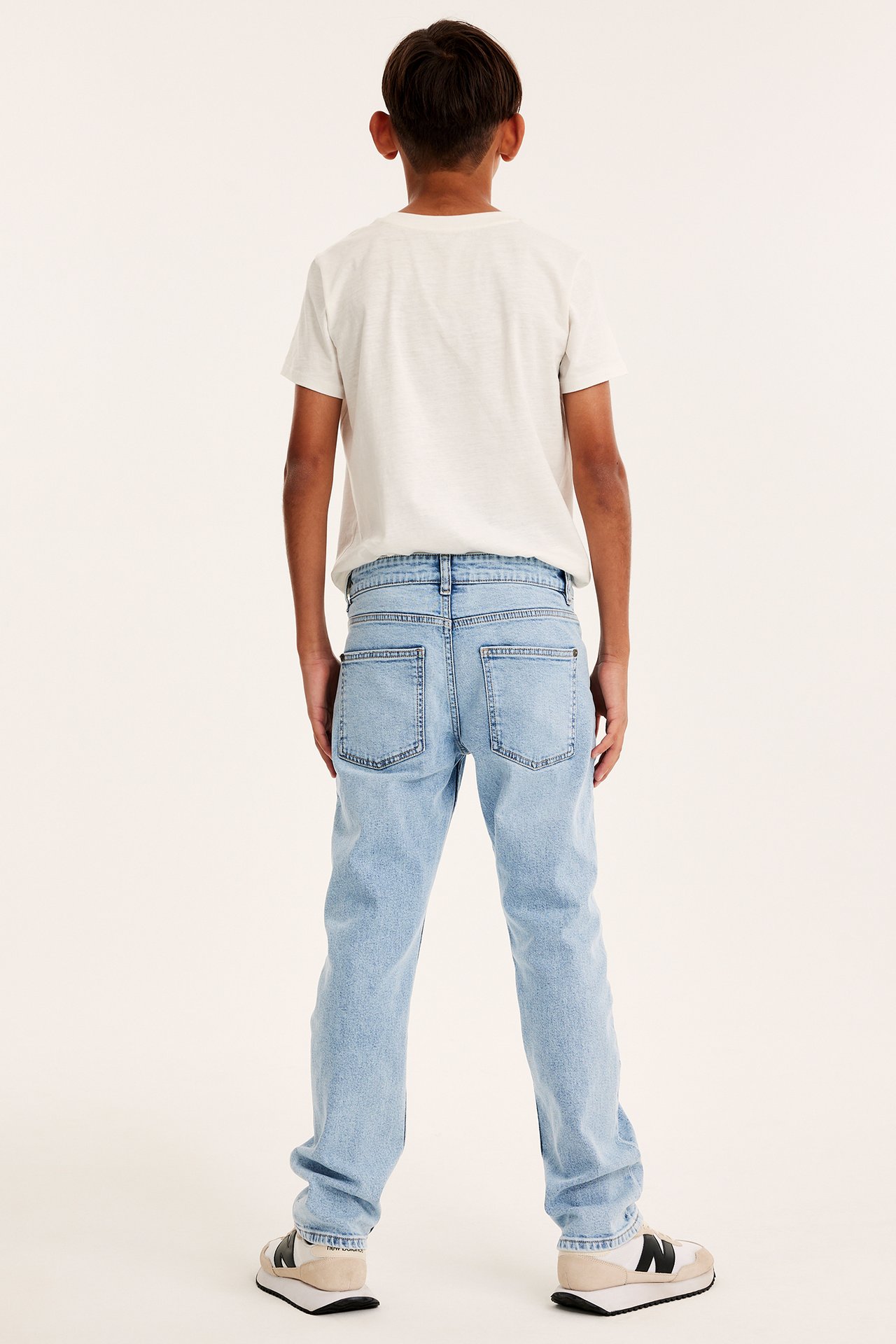 Bakifrån: ett barn i ljusblå raka jeans, en vit t-shirt och vita, svarta och beiga sneakers.