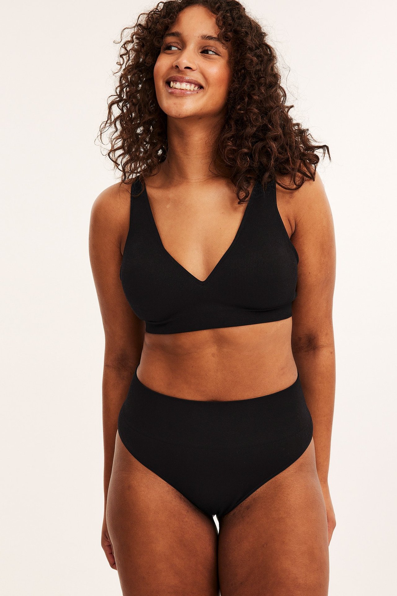 Frontbilde av kvinne i svarte ribbestrikkede sømløse brazilian-truser og matchende svart ribbet BH.