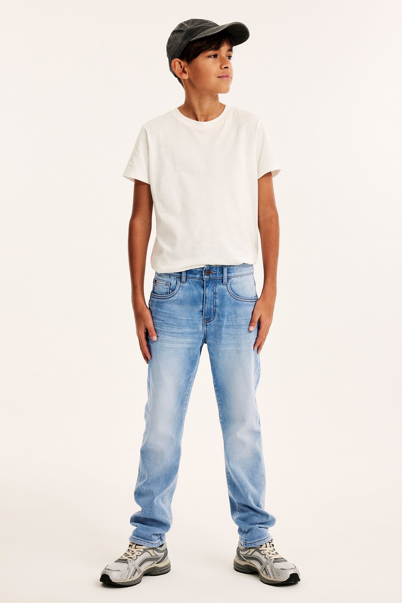 Dziecko z przodu: jasnoniebieskie jeansy relaxed jogger denim, biały t-shirt, ciemnoszara czapka, szare sneakersy.