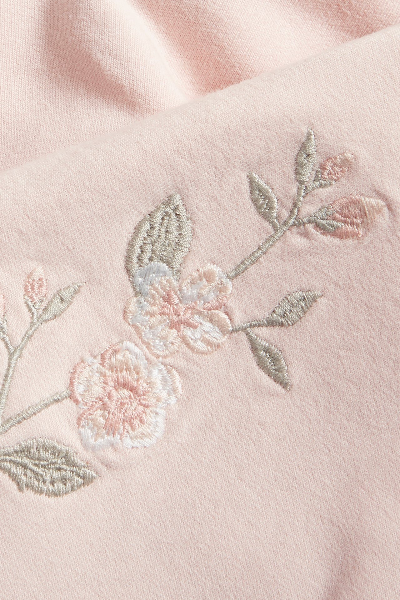 Närbild av en ljusrosa bomullshoodie med broderade ljusrosa och vita blommor samt grå blad.