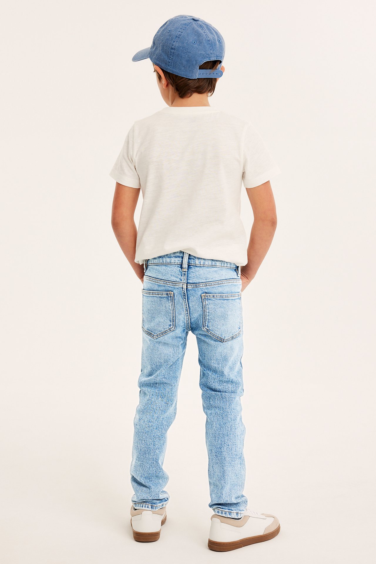 Bakifrån: ett barn i ljusblå slim-fit jeans, vit t-shirt, blå keps och vita sneakers.