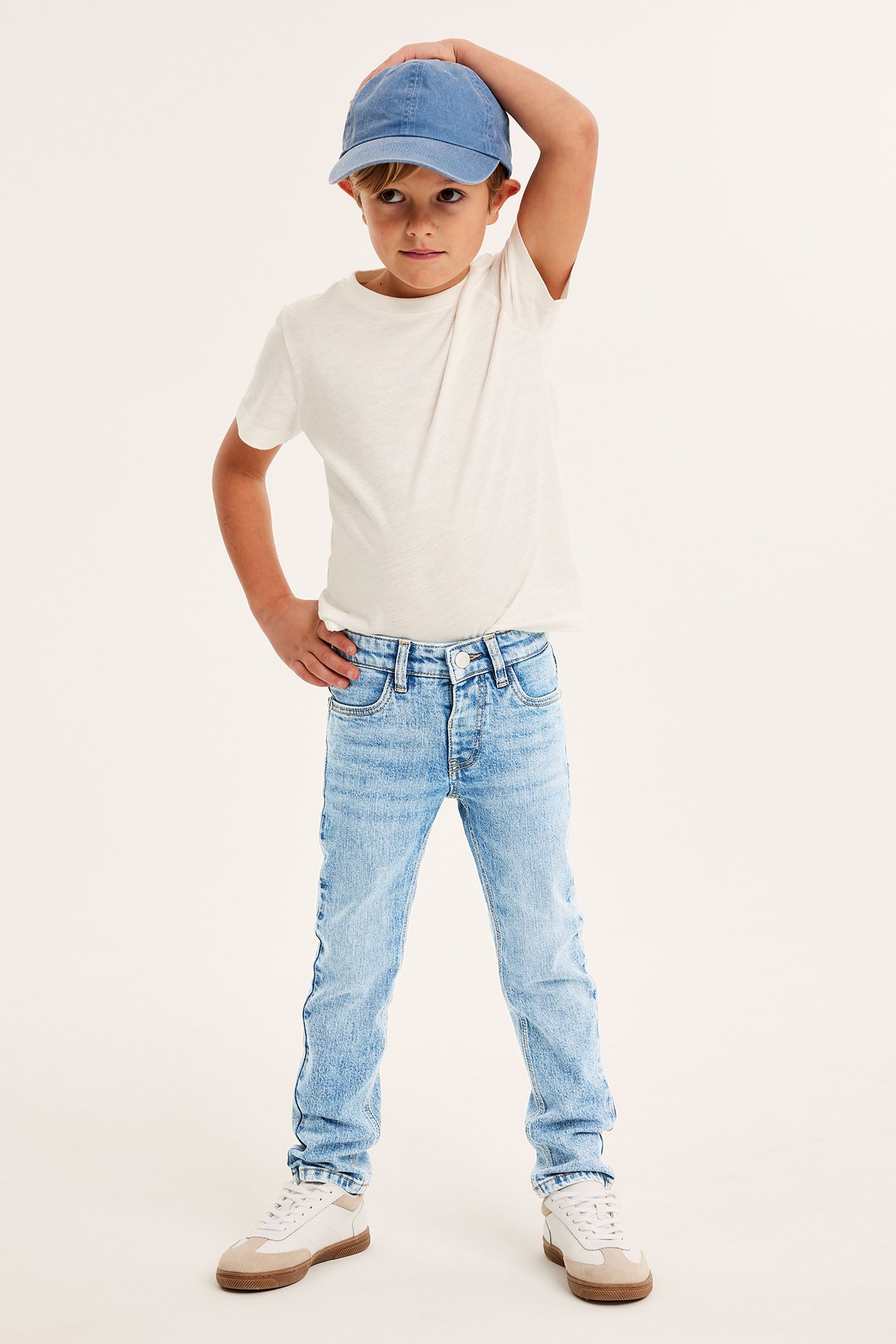 Frontvy av ett barn i ljusblå slim jeans, en ljusbeige t-shirt, blå keps och vita sneakers.
