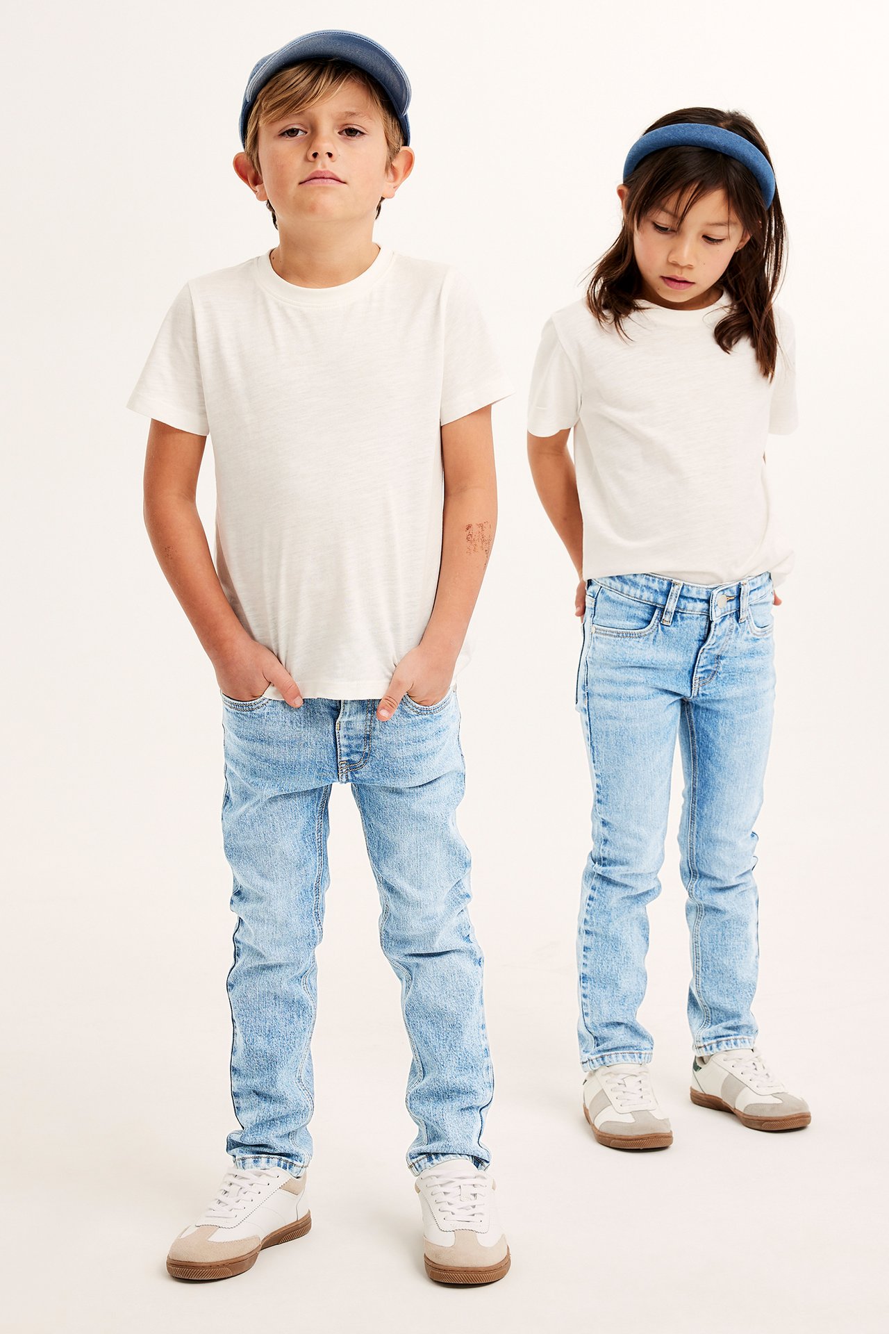 Två barn bär ljusblåa slim jeans, vita t-shirts och sneakers. Pojke med blå keps, flicka med blått hårband.