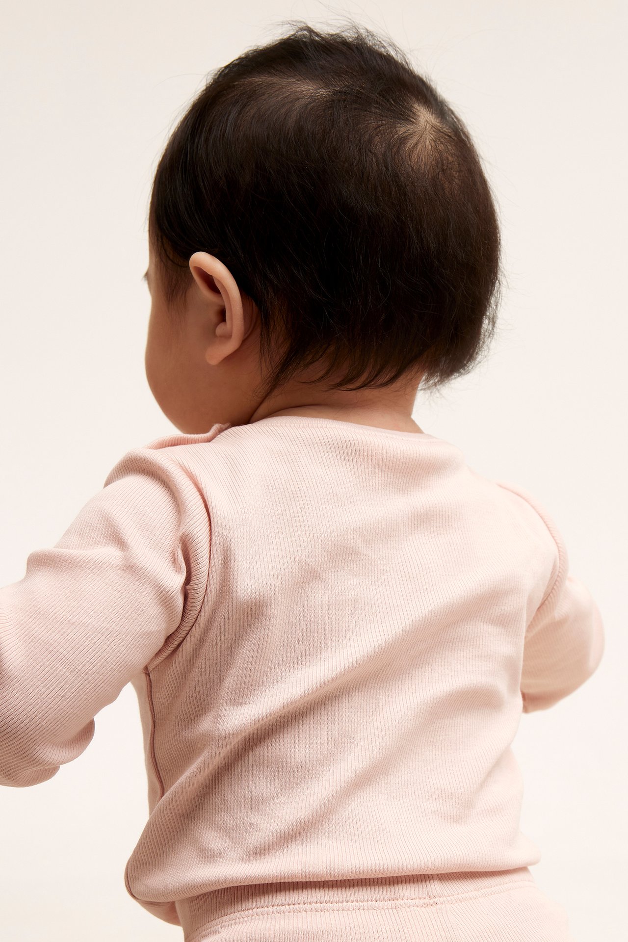 Takapuolelta katsottuna vauva vaaleanpunaisessa ribbineuloisessa pitkähihaisessa bodyssa, jossa on olkapäiden napit, ja siihen sopivissa housuissa.