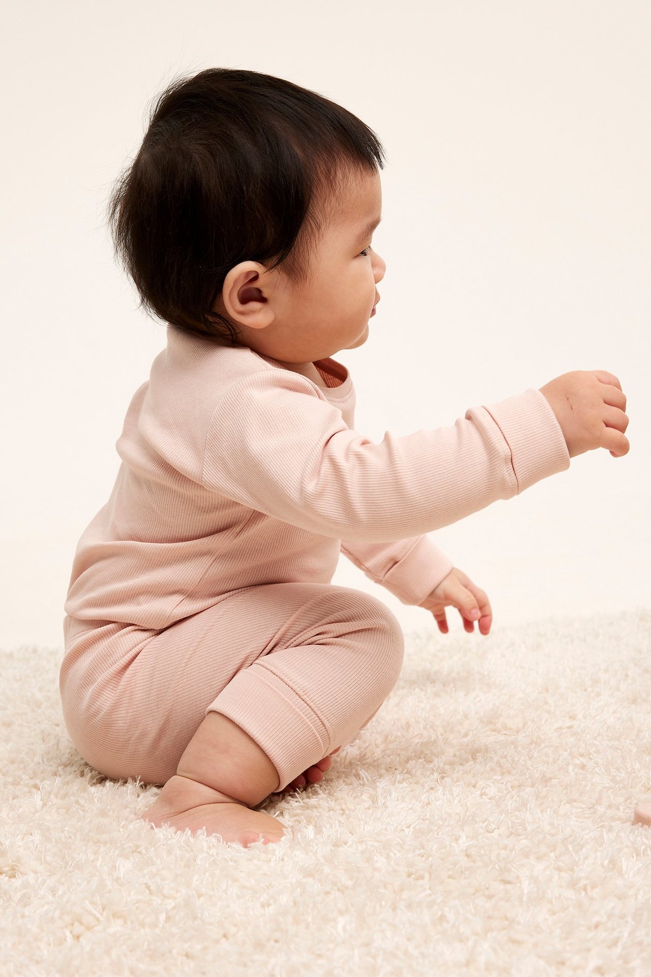 Sidovy av baby i ljusrosa ribbade leggings och matchande långärmad tröja på en matta.