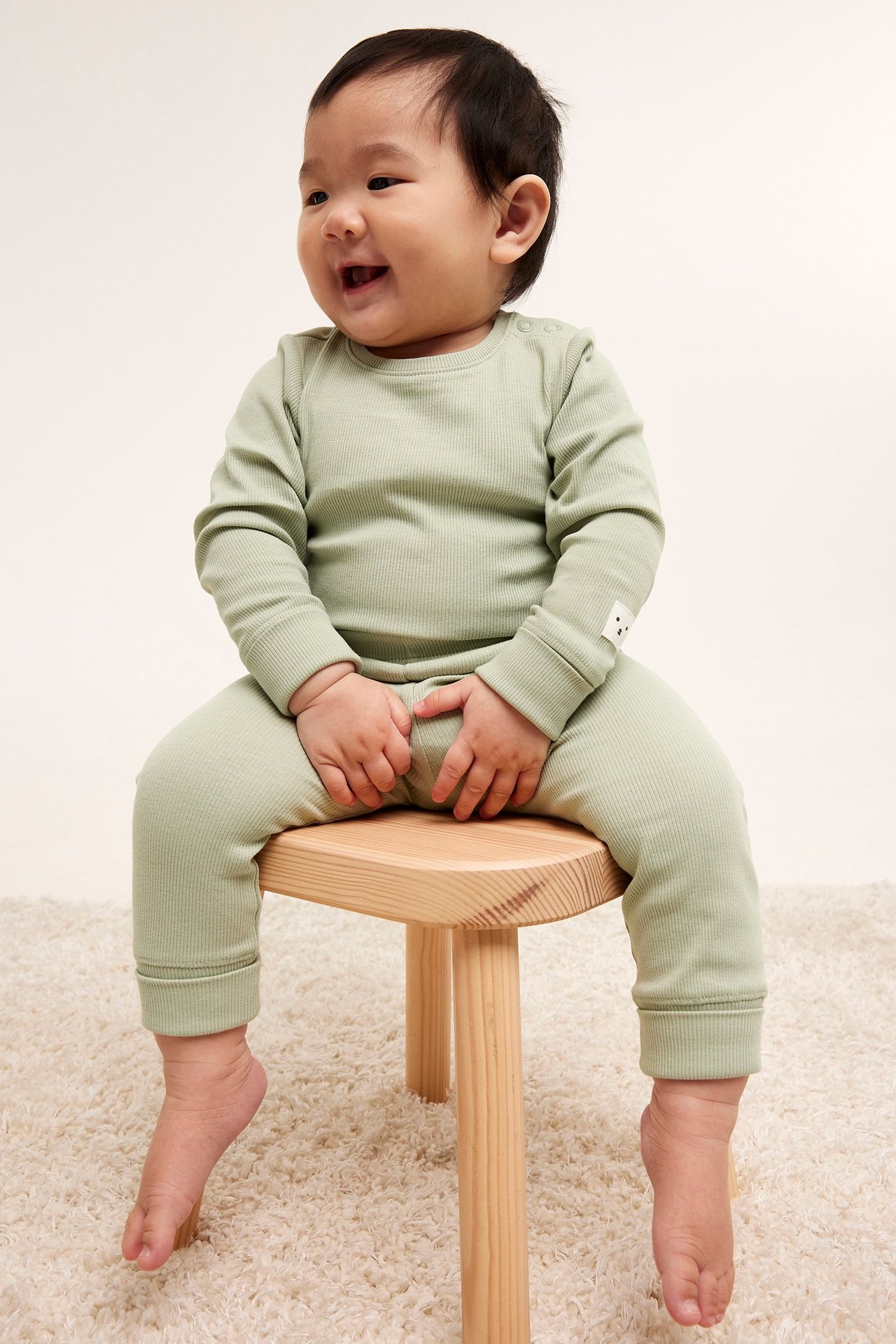 Framifrån: Leende baby i ljusgröna ribbade leggings och matchande långärmad topp, sittande på en träpall.