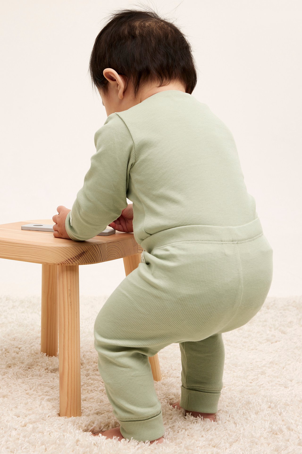 Takaa, vauva vaaleanvihreissä ribatuissa leggingseissä ja yhteensopivassa paidassa, nojaten puiseen jakkaraan.