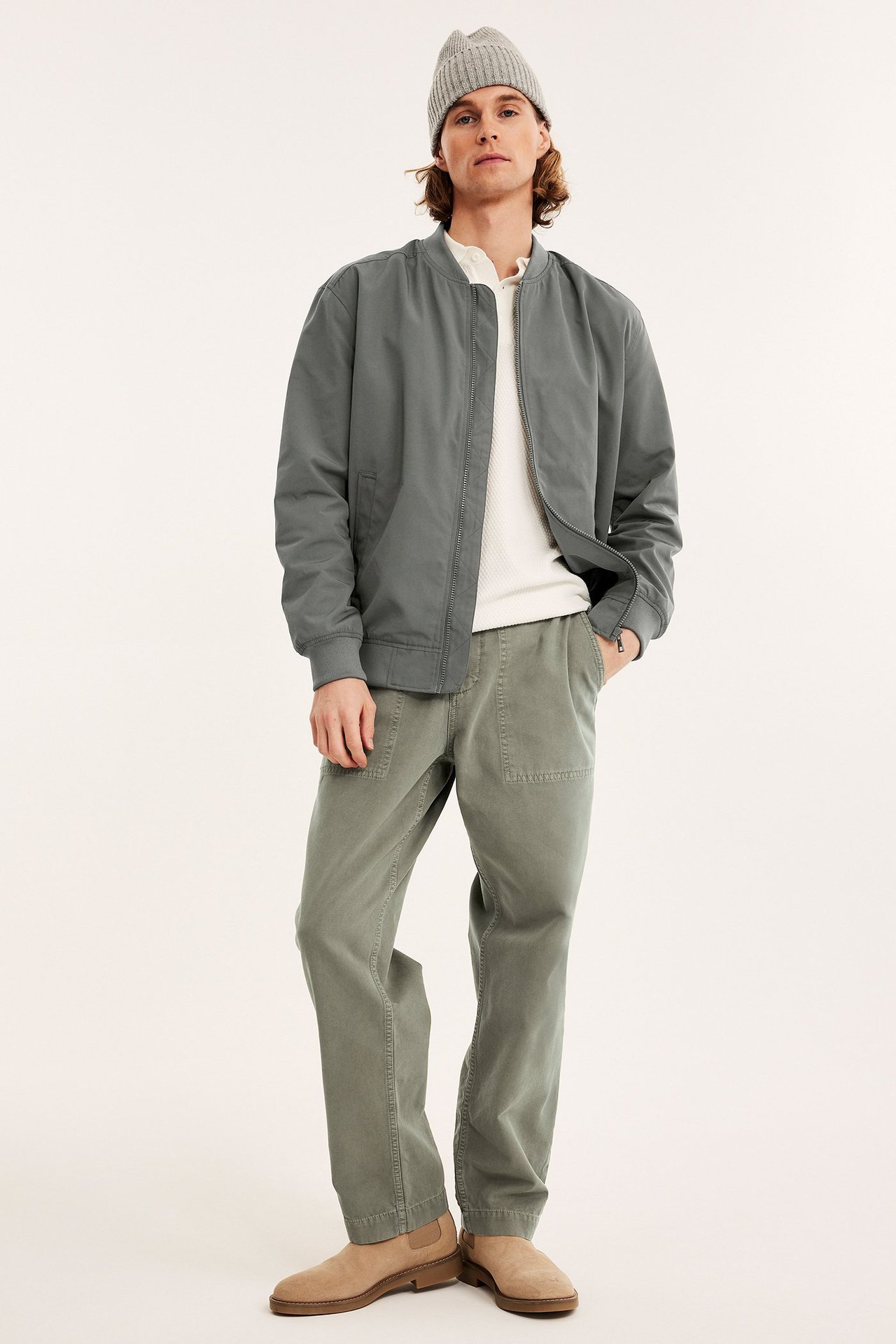 Mies tummanvihreässä bomber-takissa, valkoisessa paidassa, oliivihousuissa, harmaassa pipossa, ruskeissa kengissä.