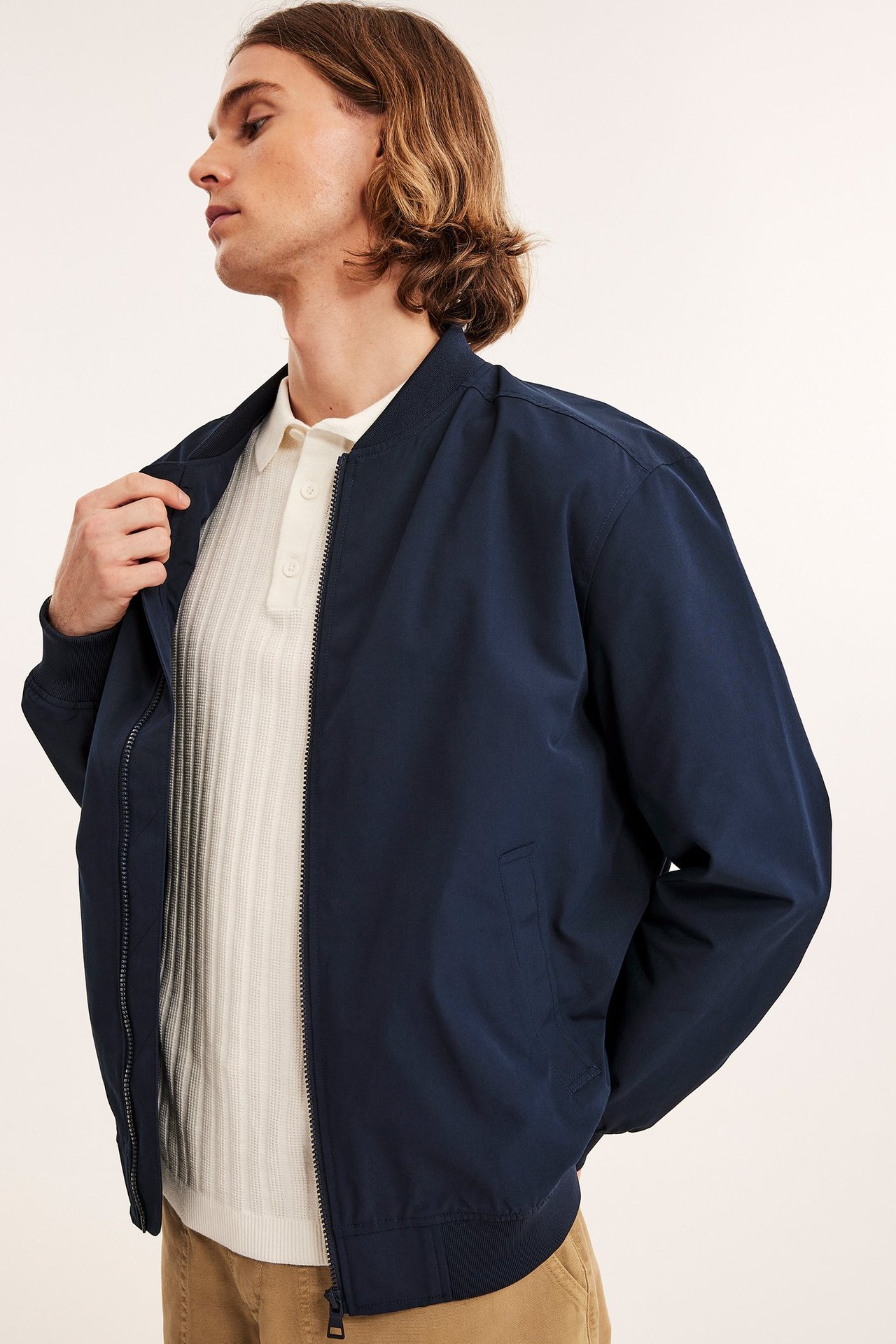 Sivukuva miehestä tummansinisessä avoimessa bomber-takissa vaalean ribbineuleisen pikeepaidan ja beigien housujen päällä.