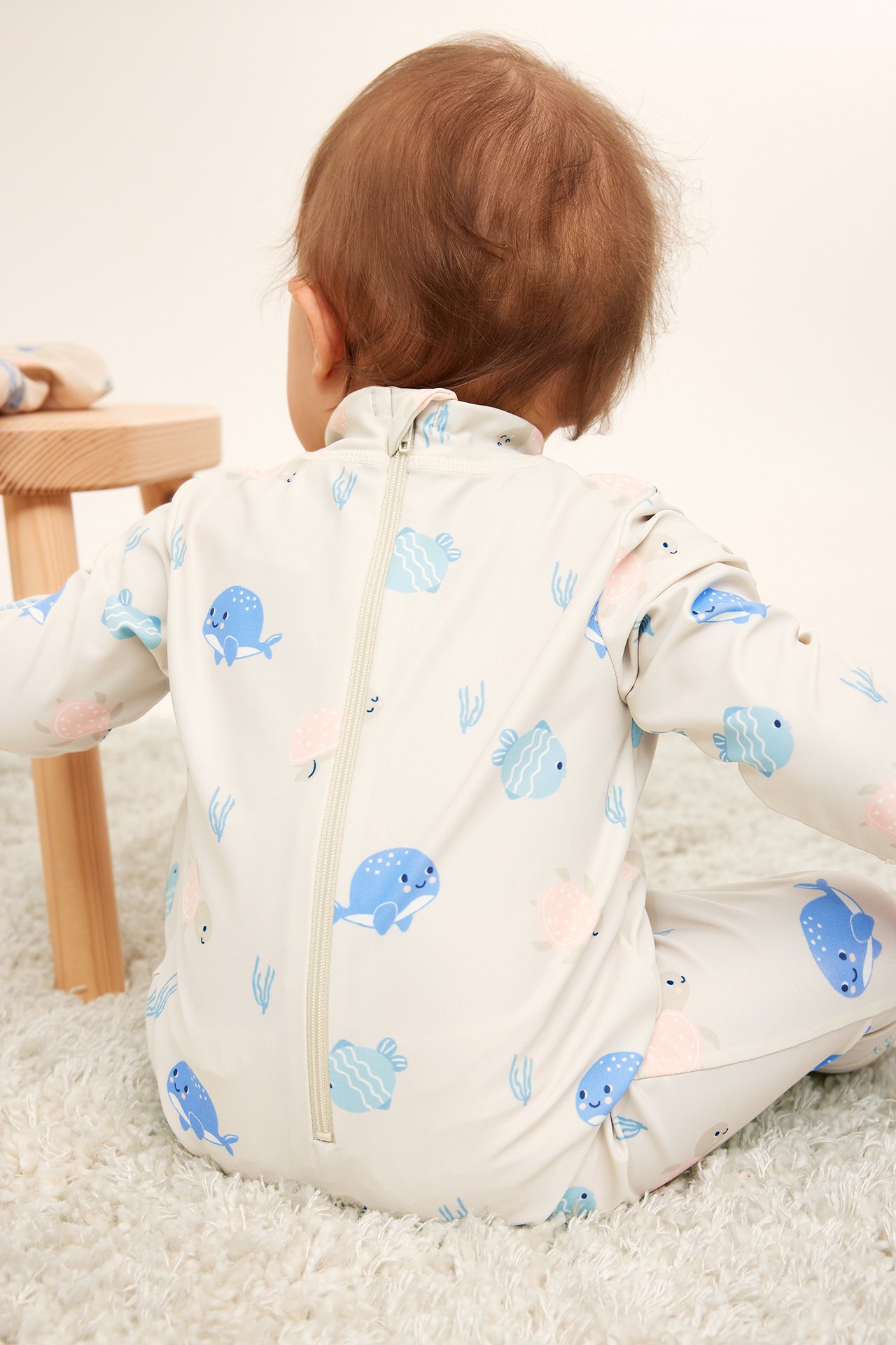 Bakifrån, baby i beige mönstrad långärmad UV-dräkt med dragkedja bak.