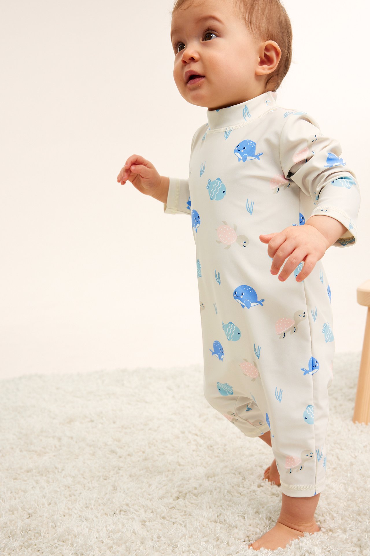 Sidovy av en baby i en beige mönstrad UV-dräkt med blå fiskar och rosa sköldpaddor.