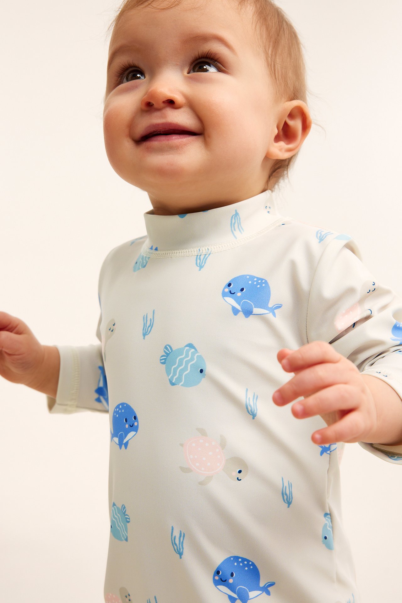 Närbild framifrån på en leende baby i en beige långärmad UV-dräkt med havsdjursmönster.