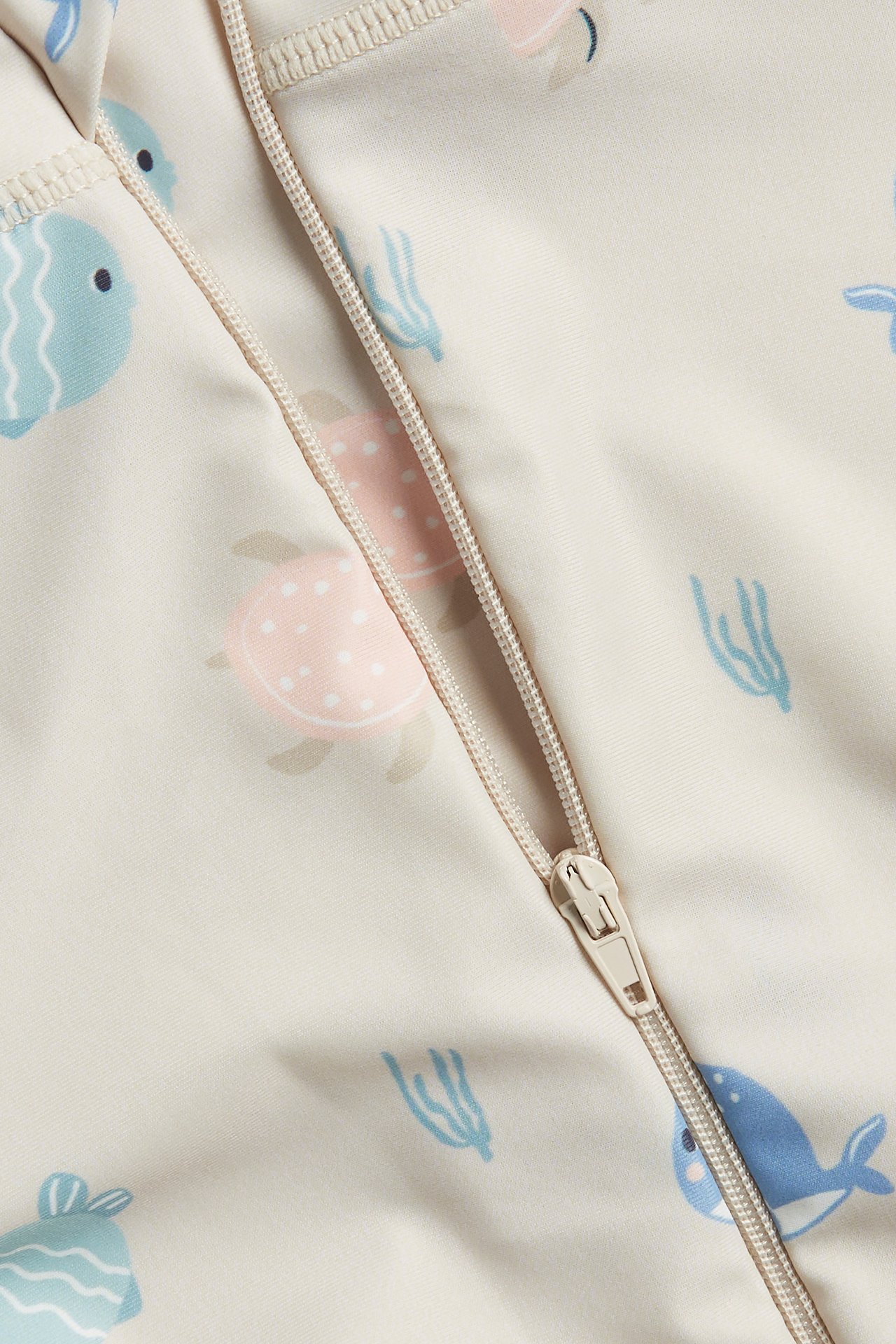 Närbild på en beige mönstrad UV-dräkt för baby med dragkedja, med blå fiskar och rosa sköldpaddor.