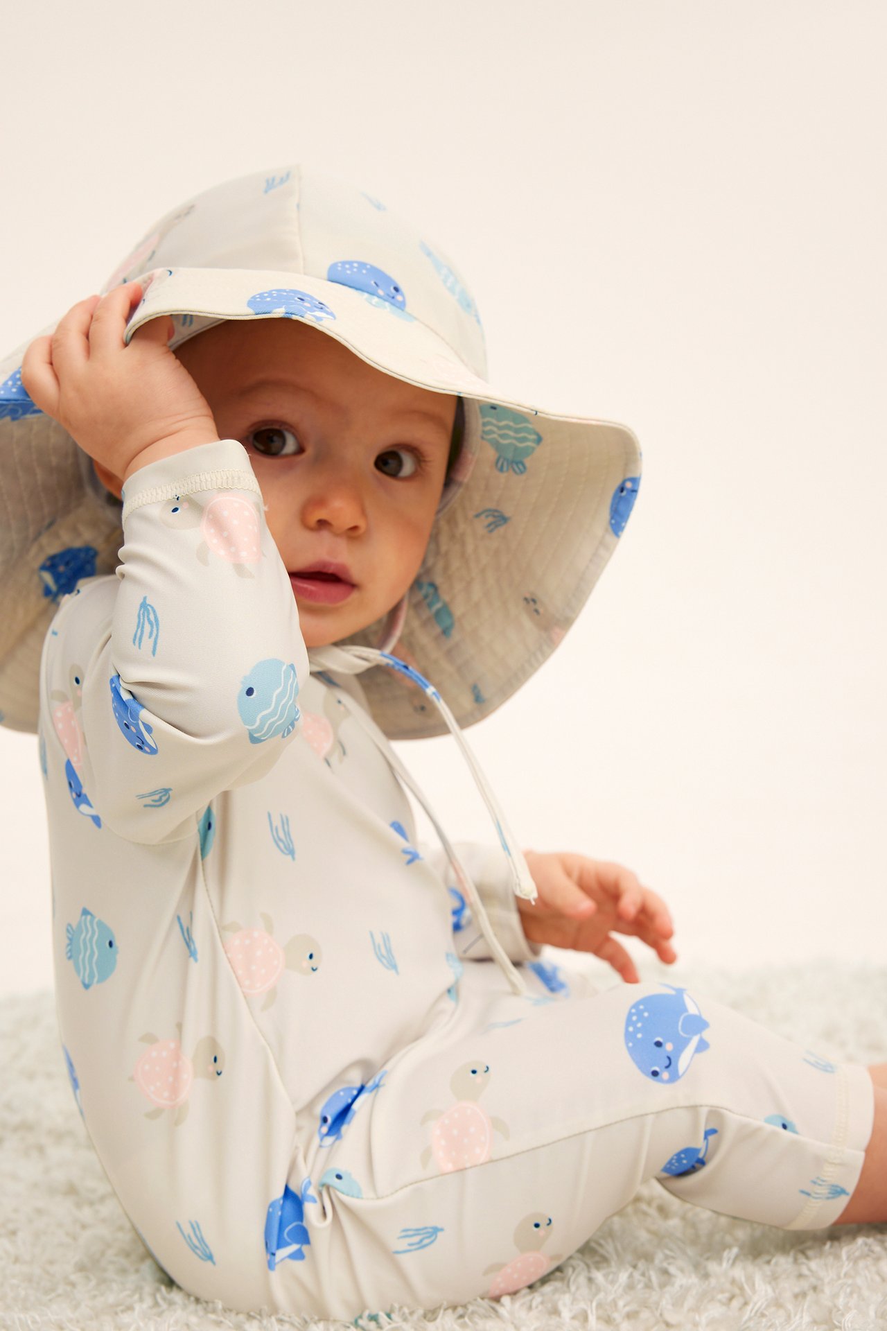 Frontvy av en baby i en beige mönstrad UV-dräkt med havsdjur och matchande solhatt.