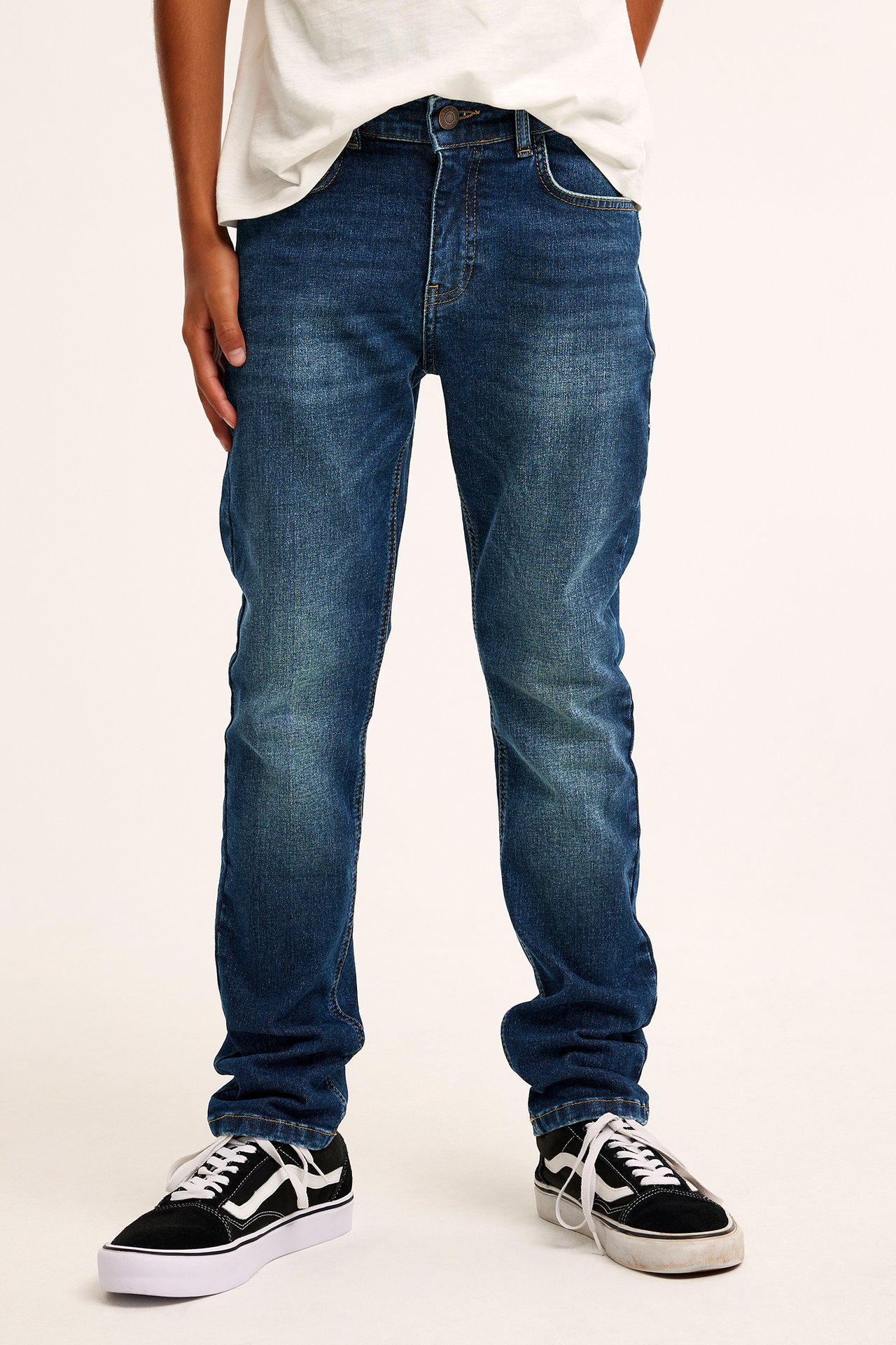 Widok z przodu: Dziecko w ciemnoniebieskich jeansach slim-fit stretch, białym t-shircie i czarno-białych trampkach.