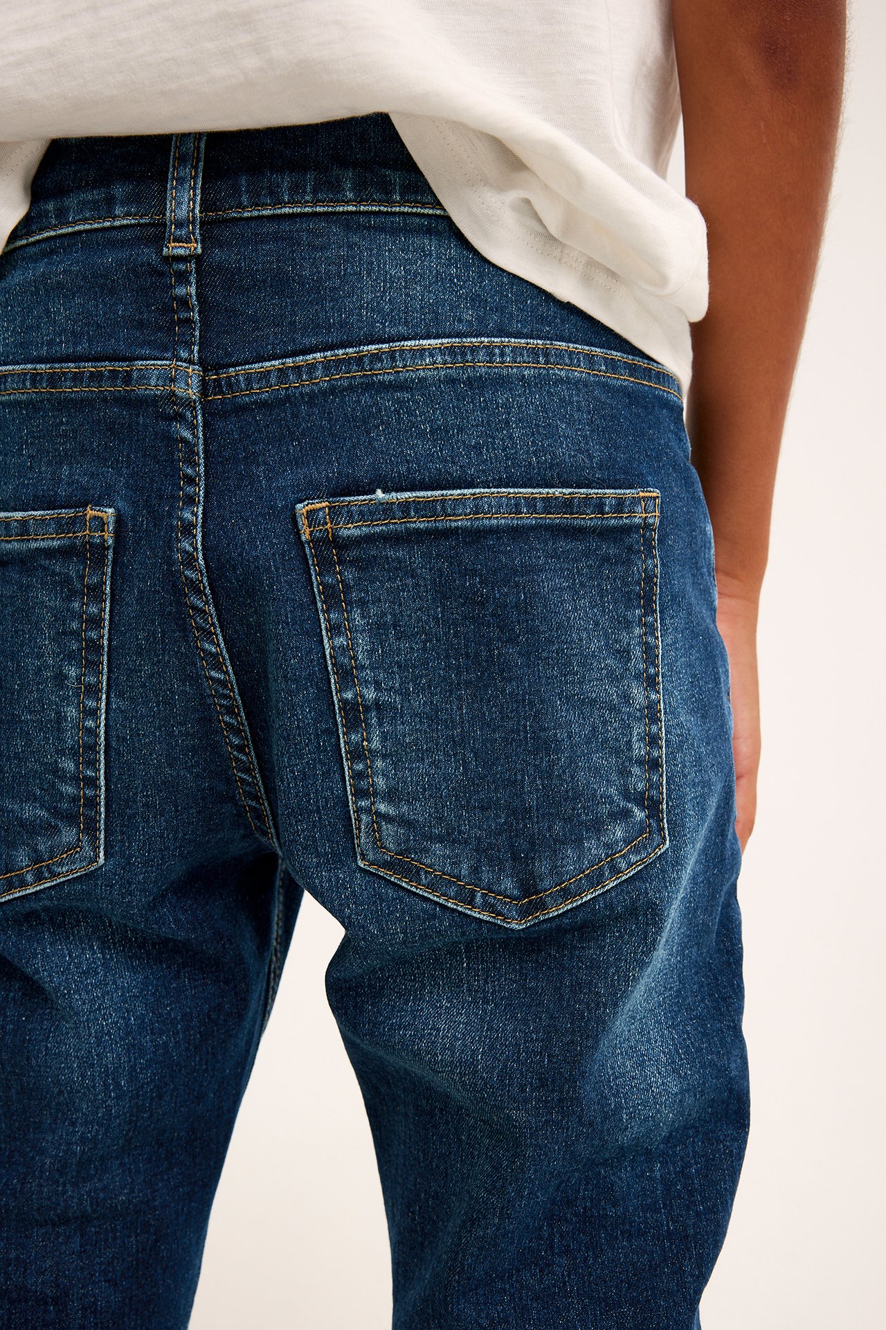 Widok z tyłu: dziecko w ciemnoniebieskich slim-fit jeansach z żółtymi szwami i białym t-shirtem.