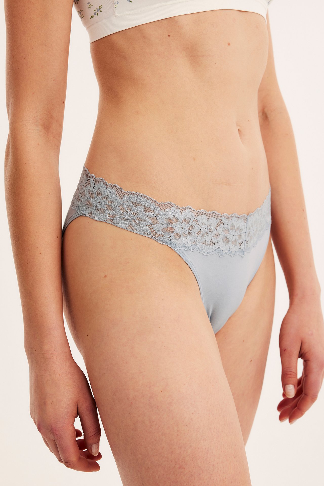 Sidebilde av en kvinne i lyseblå brazilian-truse med dekorativ blondekant i midjen og hvit blomstret BH.