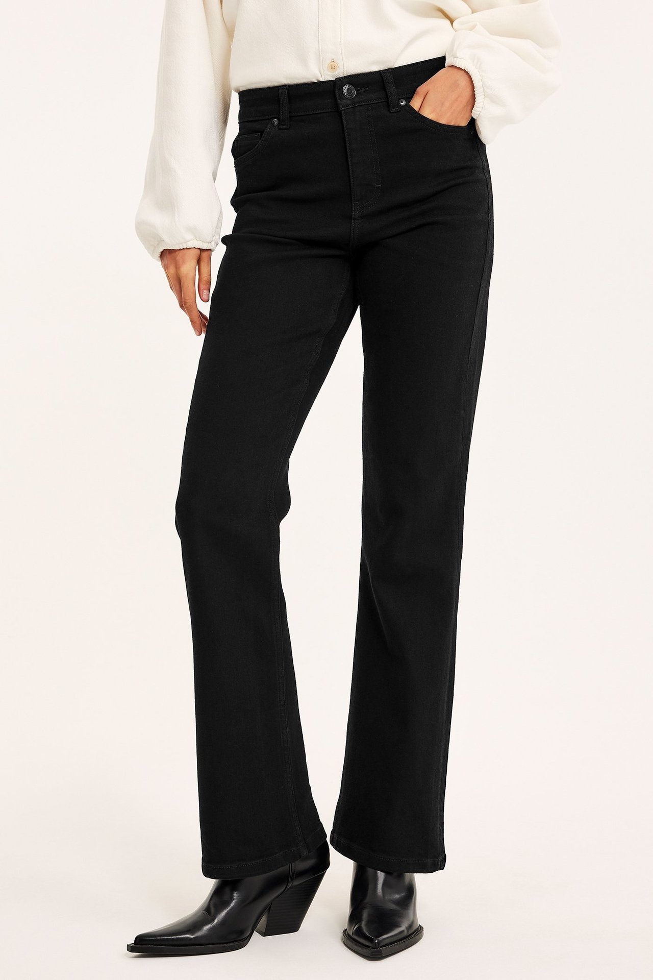 Frontvy av en kvinna i svarta flare jeans med normal midja, en ljus blus och svarta stövlar.