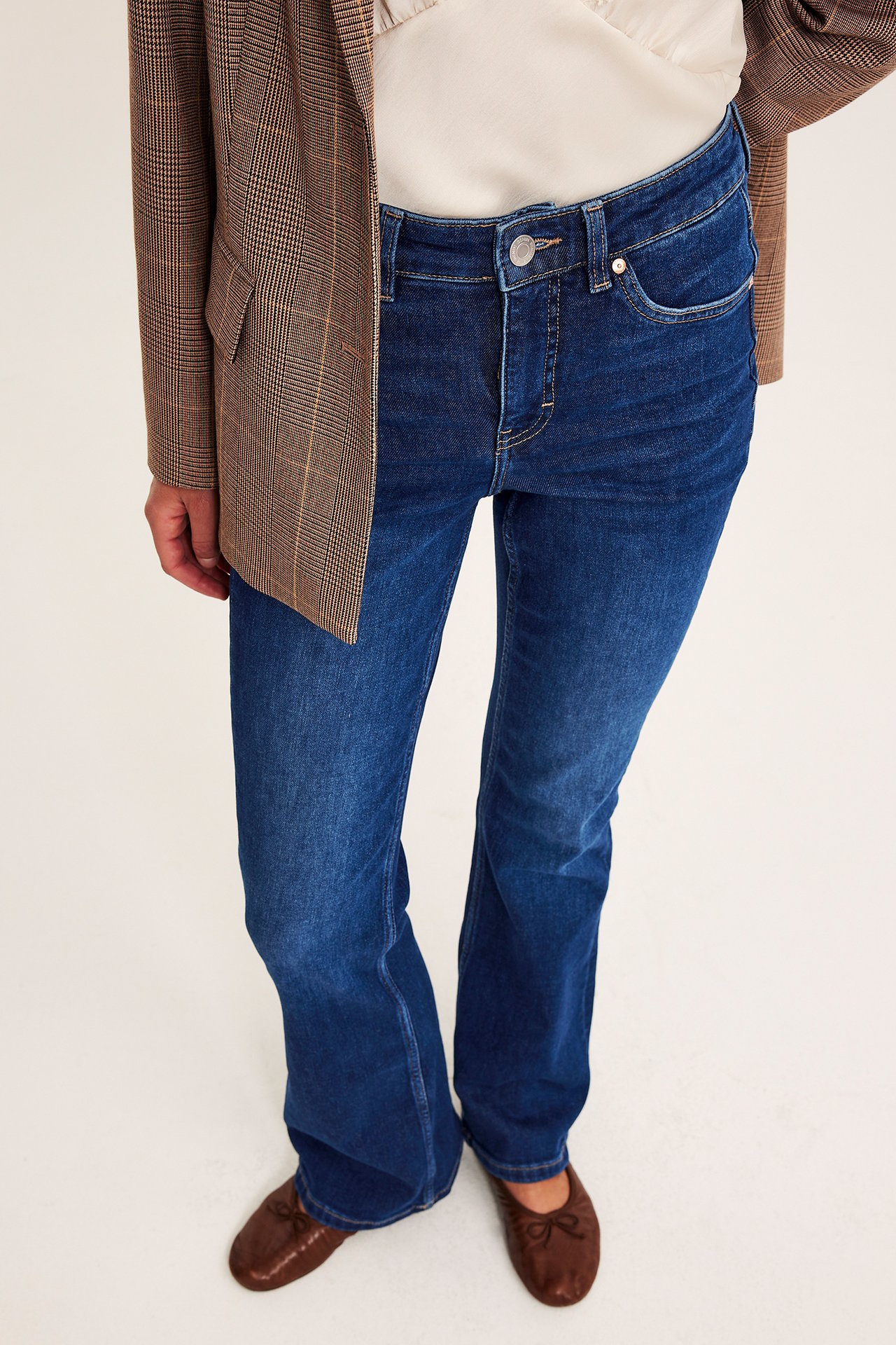 Framifrån: Kvinna i mörkblå bootcut-jeans, brunrutig kavaj och bruna ballerinaskor.