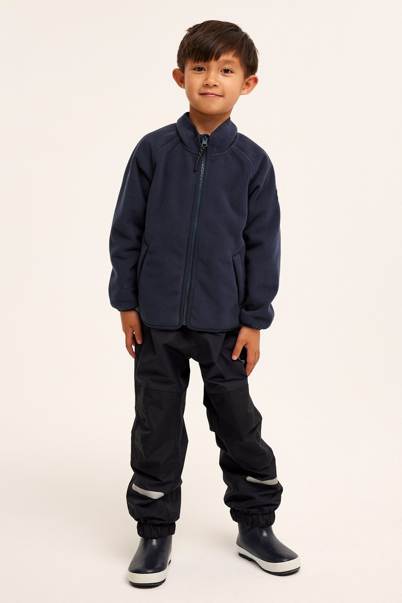 Edestä: lapsi tummansinisessä fleecetakissa, tummansinisissä housuissa heijastimilla ja kumisaappaissa.