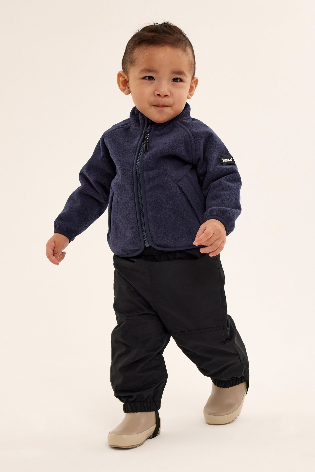 Widok z przodu dziecka w ciemnoniebieskiej polarowej kurtce z zamkiem, czarnych spodniach i jasnobrązowych butach.