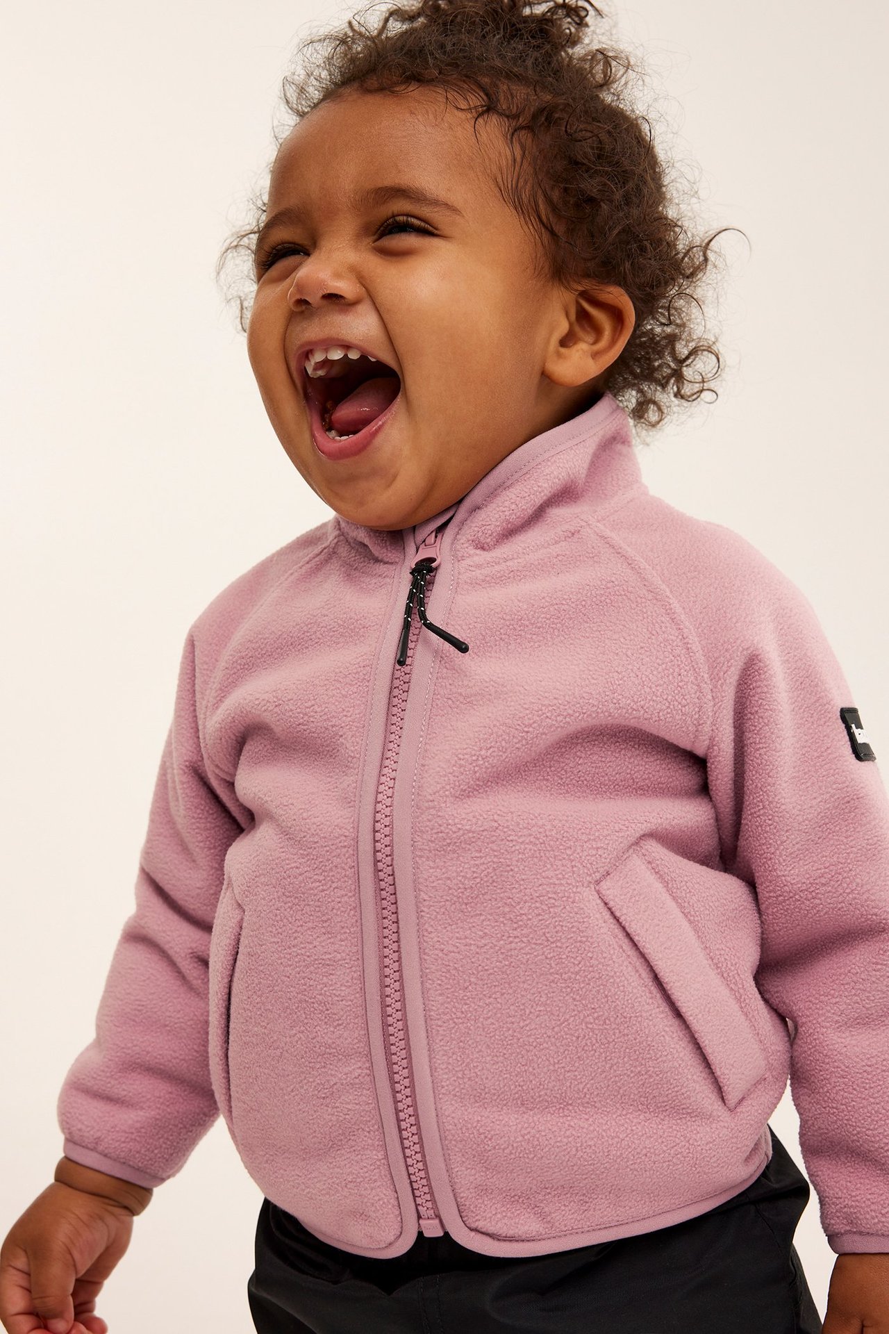 Zbliżenie na śmiejące się dziecko w różowej polarowej kurtce z zamkiem i kieszeniami, na czarnych spodniach.