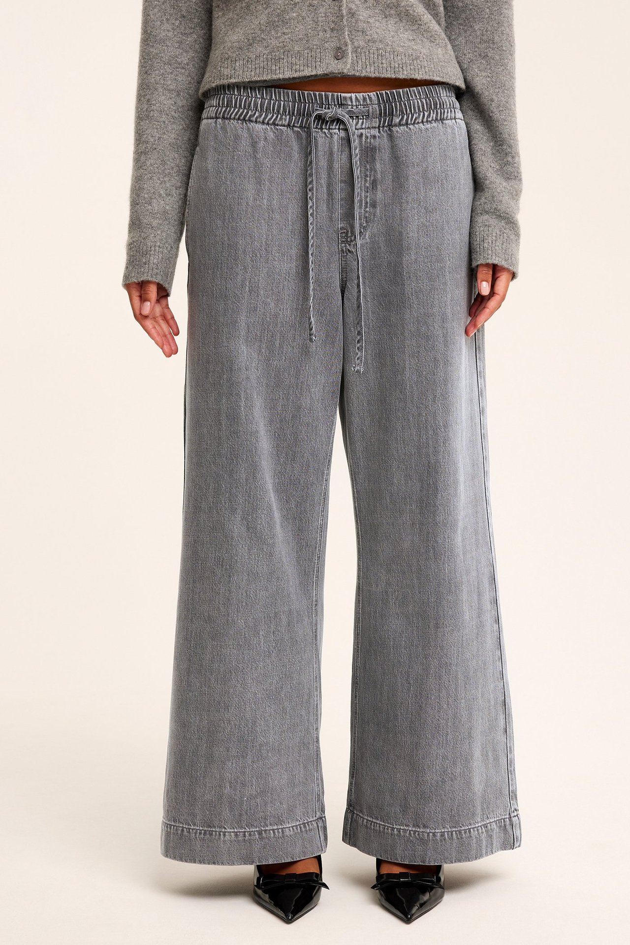 Framifrån: Kvinna i vida grå denimjeans med dragsko i midjan, grå kofta och svarta skor.