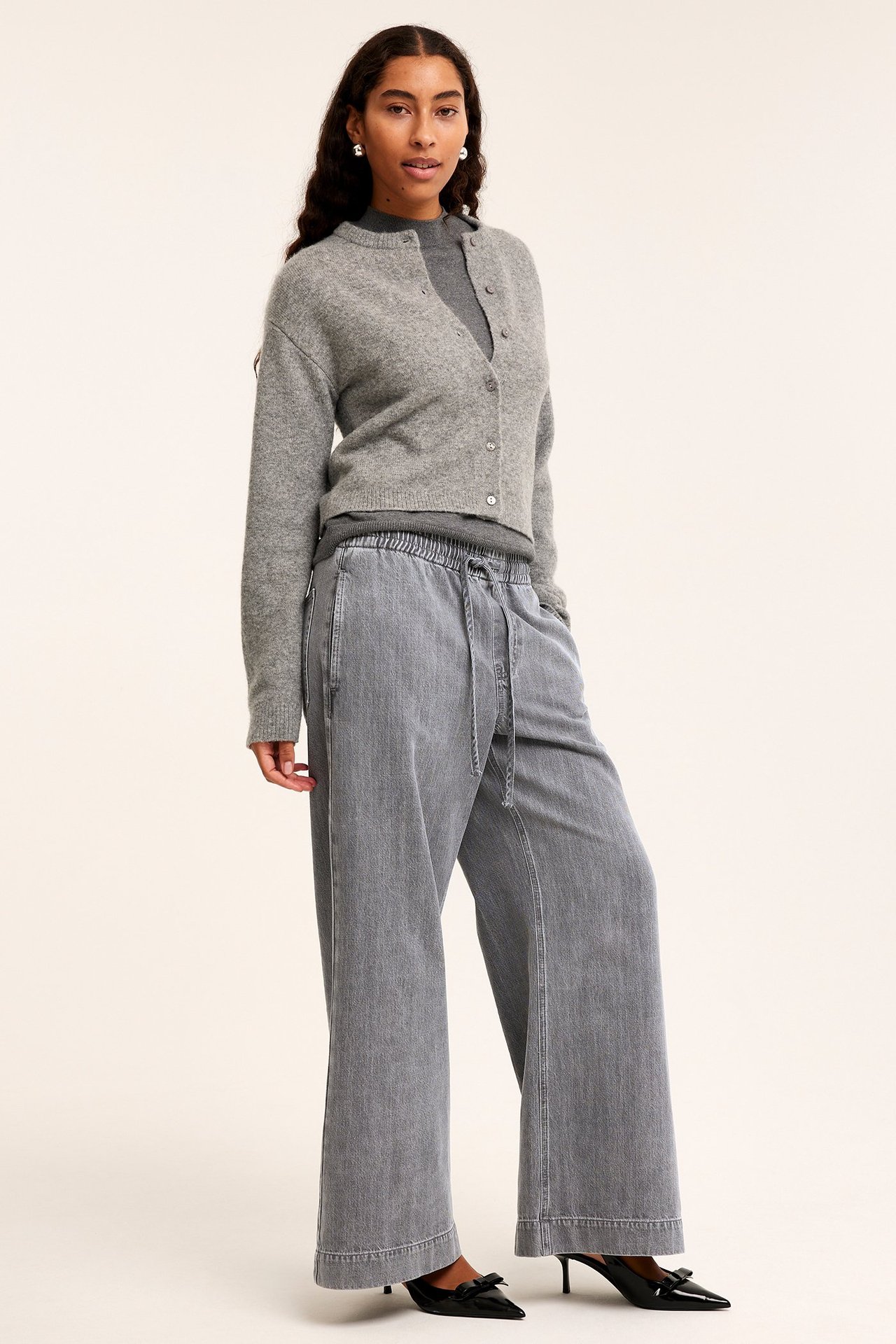 Framifrån: Kvinna i grå denim vida pull-on jeans, grå kofta, polotröja och svarta klackar.