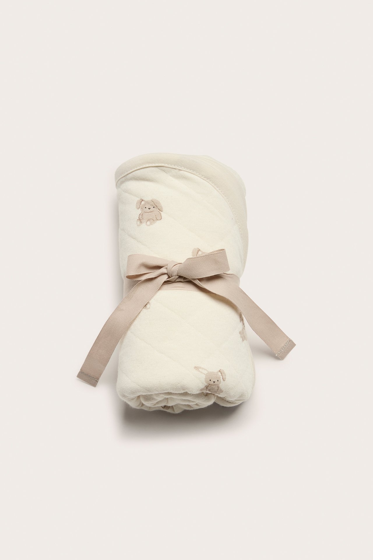 Forfra, et rullet offwhite vattert babyteppe med kaninmønster, bundet med et beige bånd.