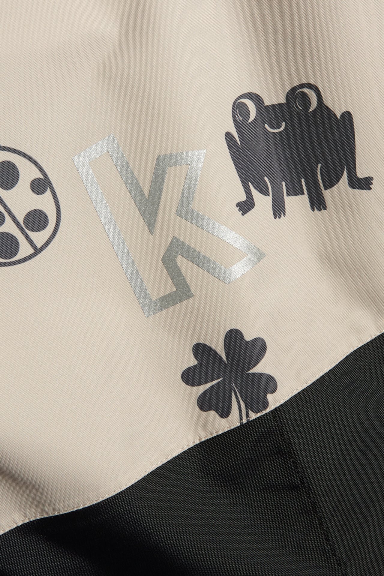 Lähikuva beigestä ja mustasta kuvioidusta Kaxs-kuorihaalarista, jossa on sammakko-, leppäkerttu-, apila- ja K-kirjainkuvioita.
