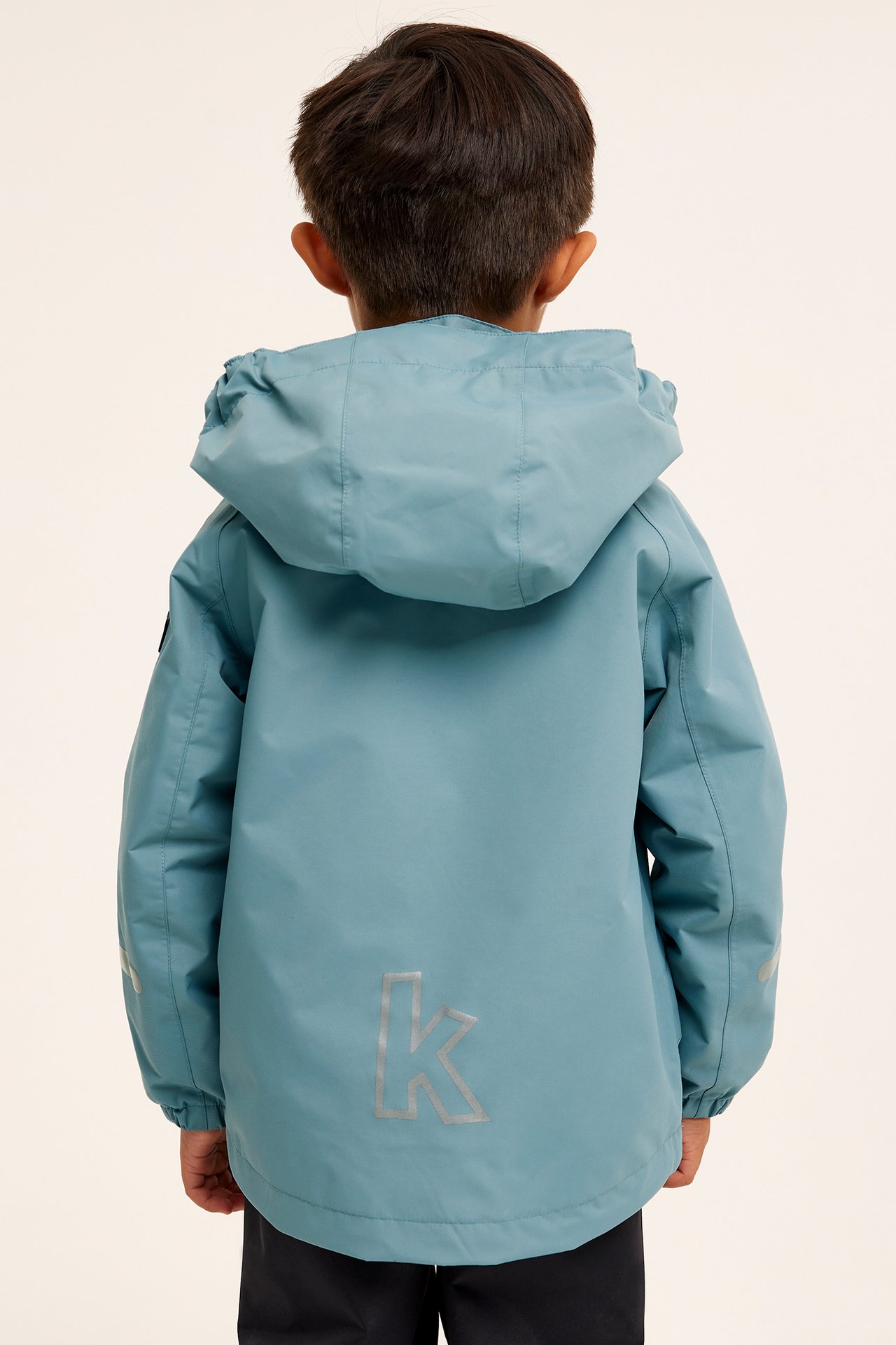Widok z tyłu dziecka w niebieskiej kurtce softshell z kapturem, odblaskami i ciemnych spodniach.