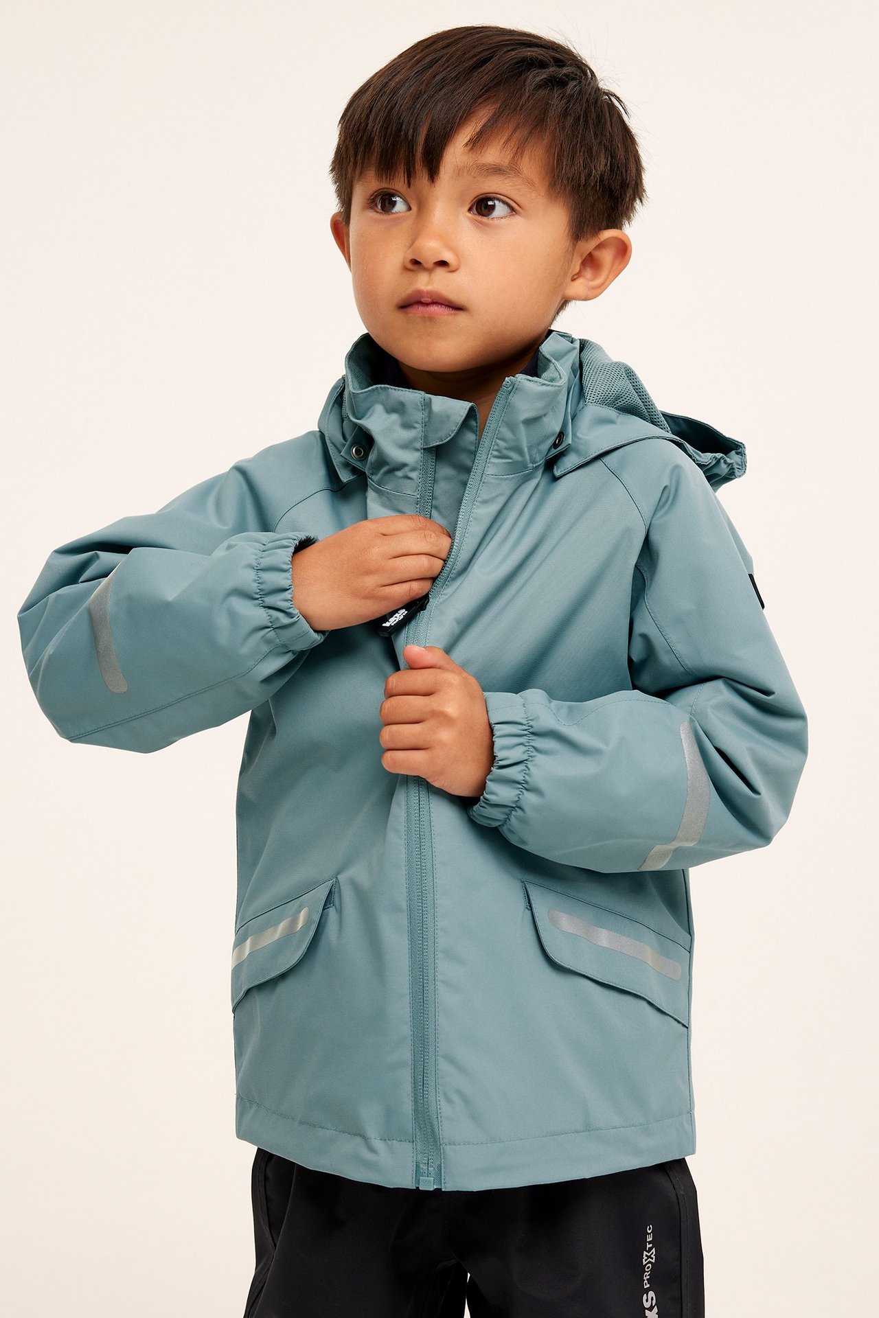 Frontvy av ett barn som drar upp en blå vattentät skaljacka med reflexdetaljer och svarta byxor.