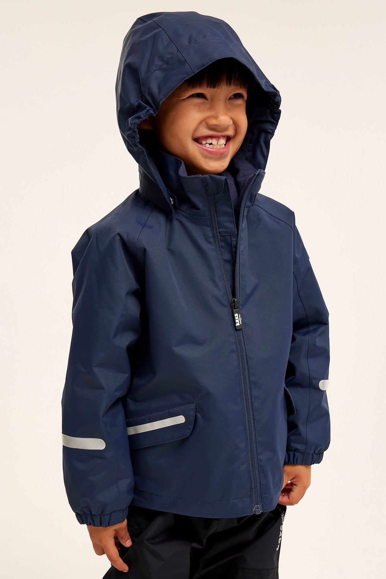 Widok z przodu uśmiechniętego dziecka w granatowej wodoodpornej kurtce z kapturem i odblaskami.