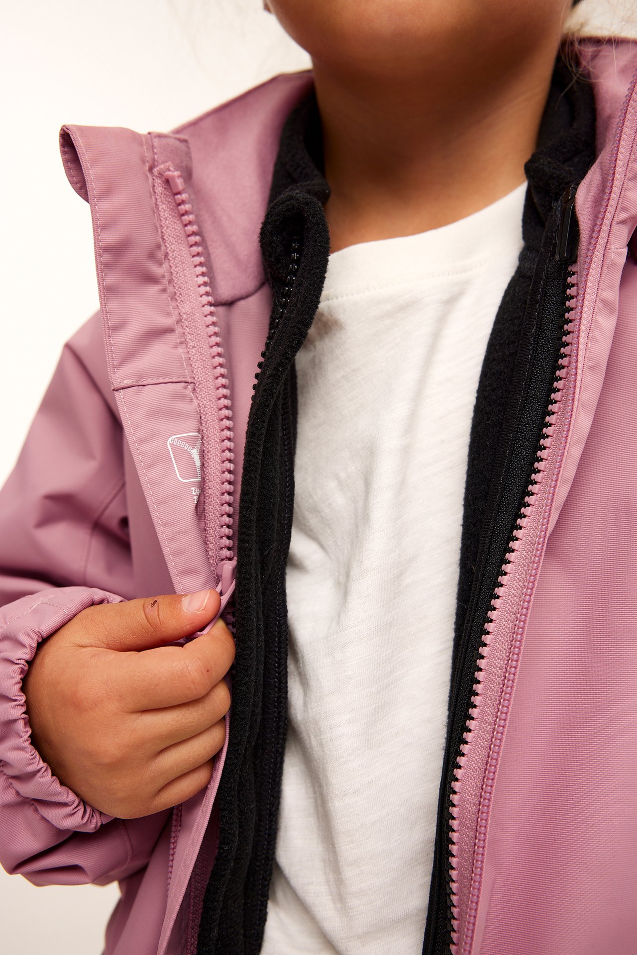 Närbild framifrån på ett barn i en rosa skaljacka över en svart fleece och vit t-shirt.