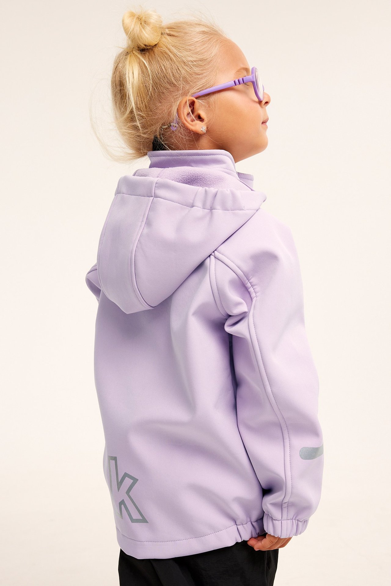 Sidovy av ett barn i lila softshelljacka med huva, svarta byxor och lila glasögon.