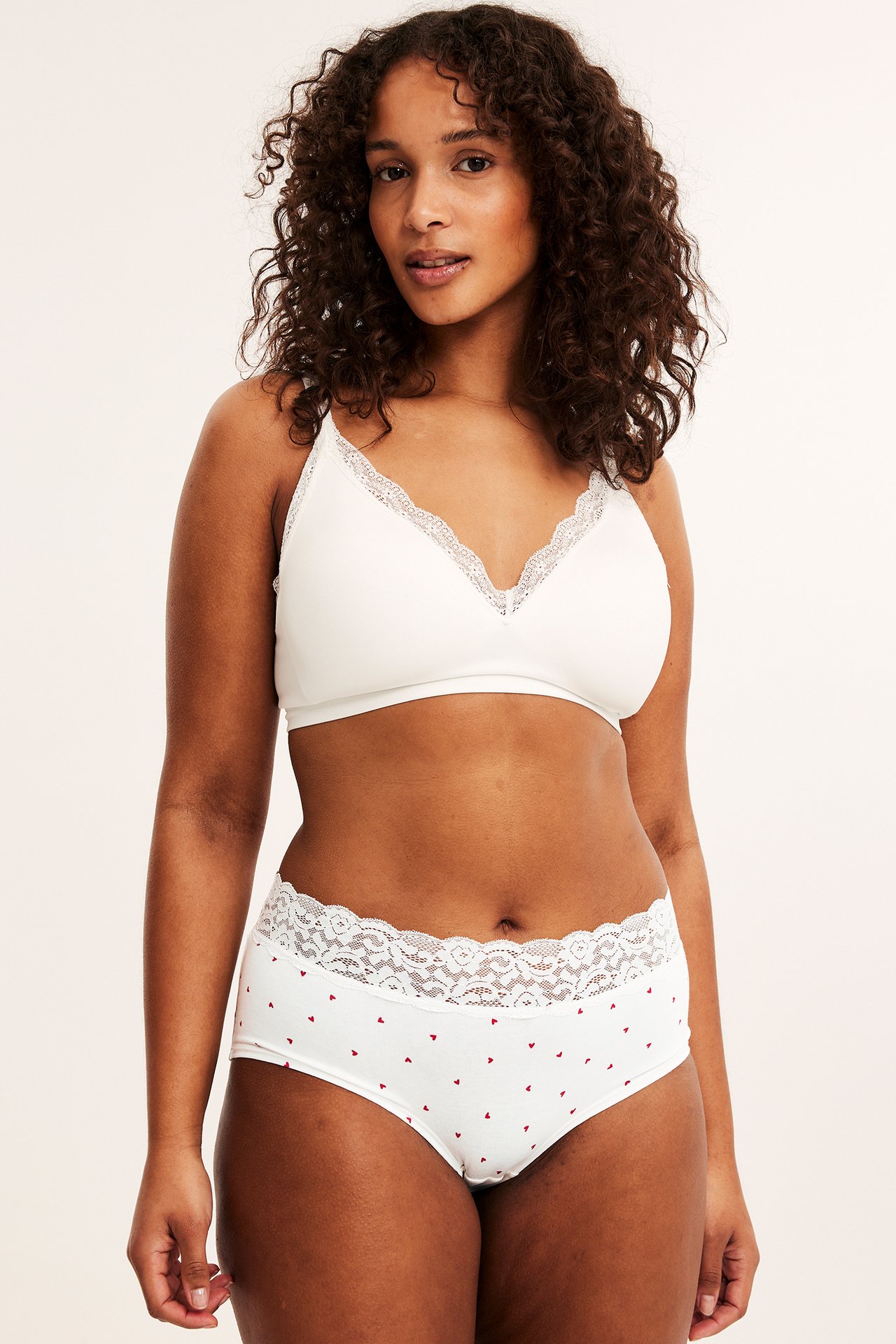 Framifrån, kvinna i vita bomullshipstertrosor med röda hjärtan och vit spets, samt en vit bralette.