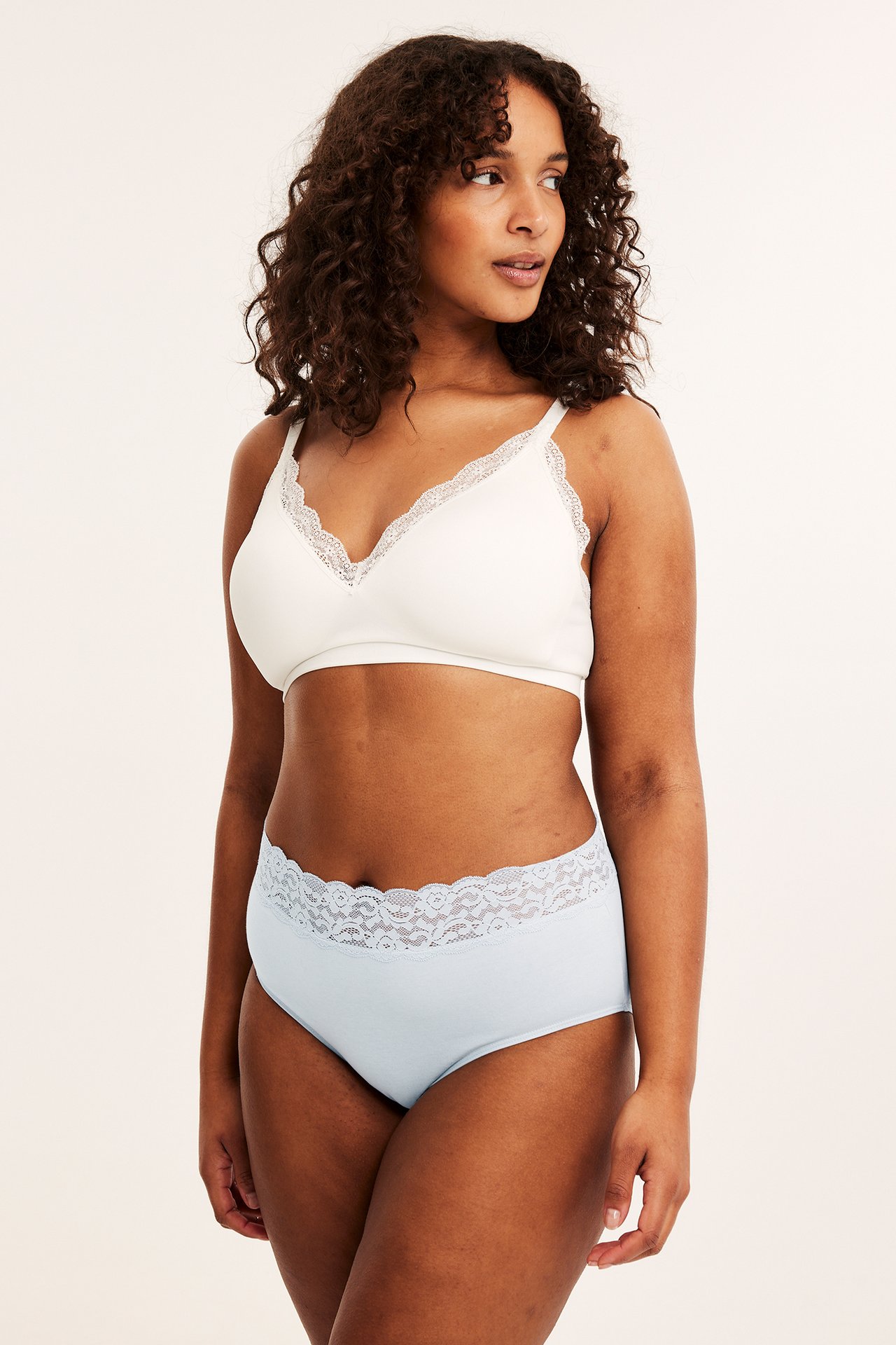Nainen edestäpäin yllään vaaleansiniset mid-brief alushousut pitsivyötäröllä ja valkoiset pitsireunaiset rintaliivit.