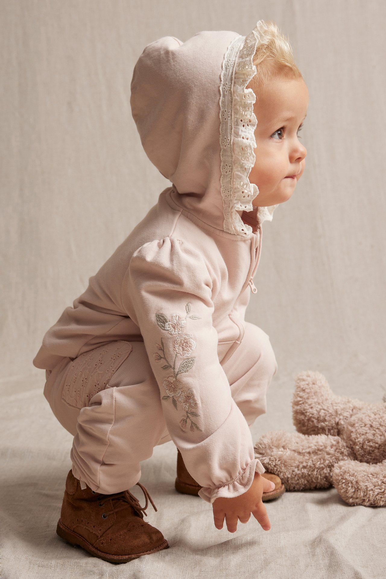 Sidovy: Baby i ljusrosa hoodie med blommig brodyr, spetskantad huva, matchande byxor och bruna kängor.