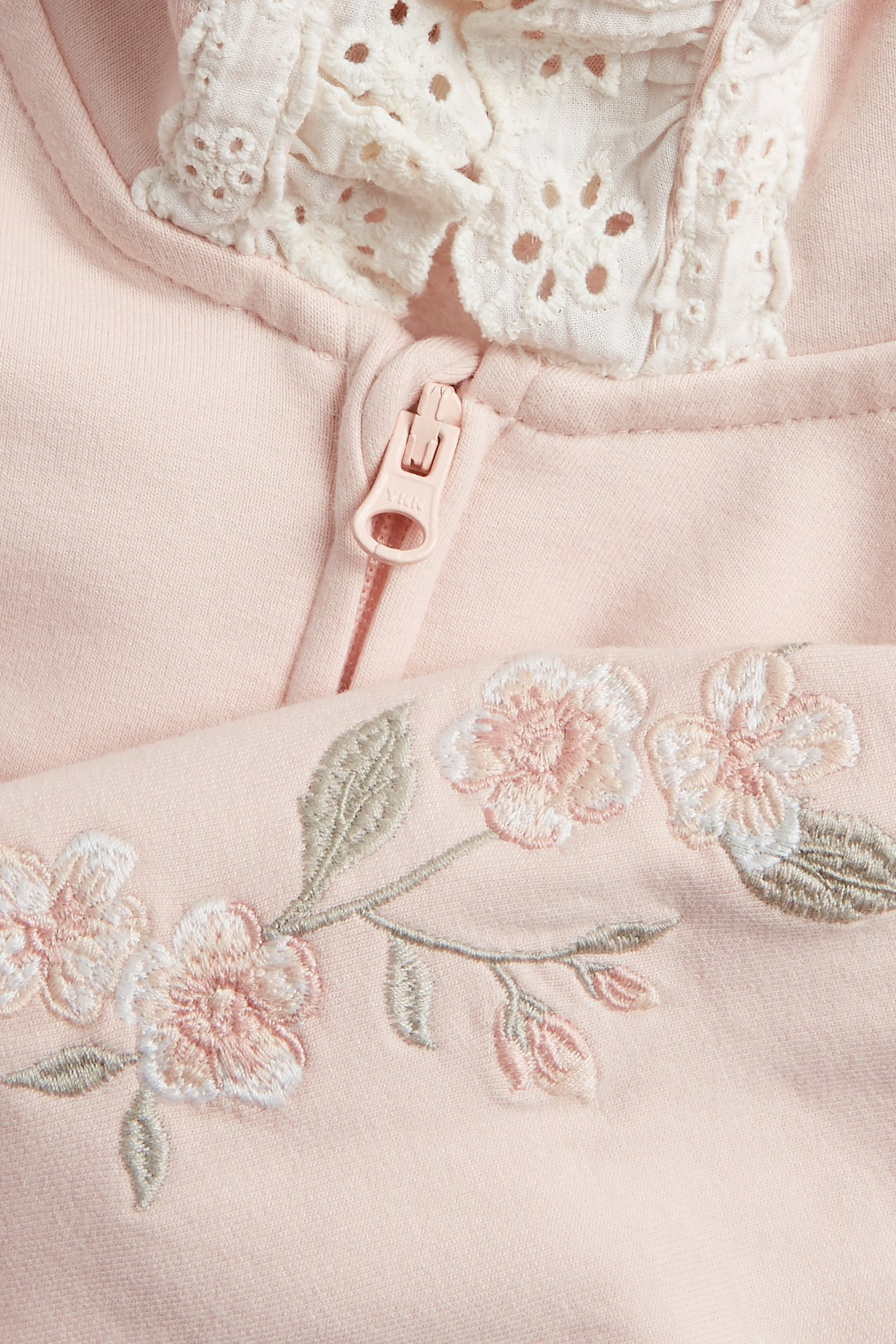 Närbild av en ljusrosa hoodie med dragkedja, blommig brodyr och en vit spetskrage.