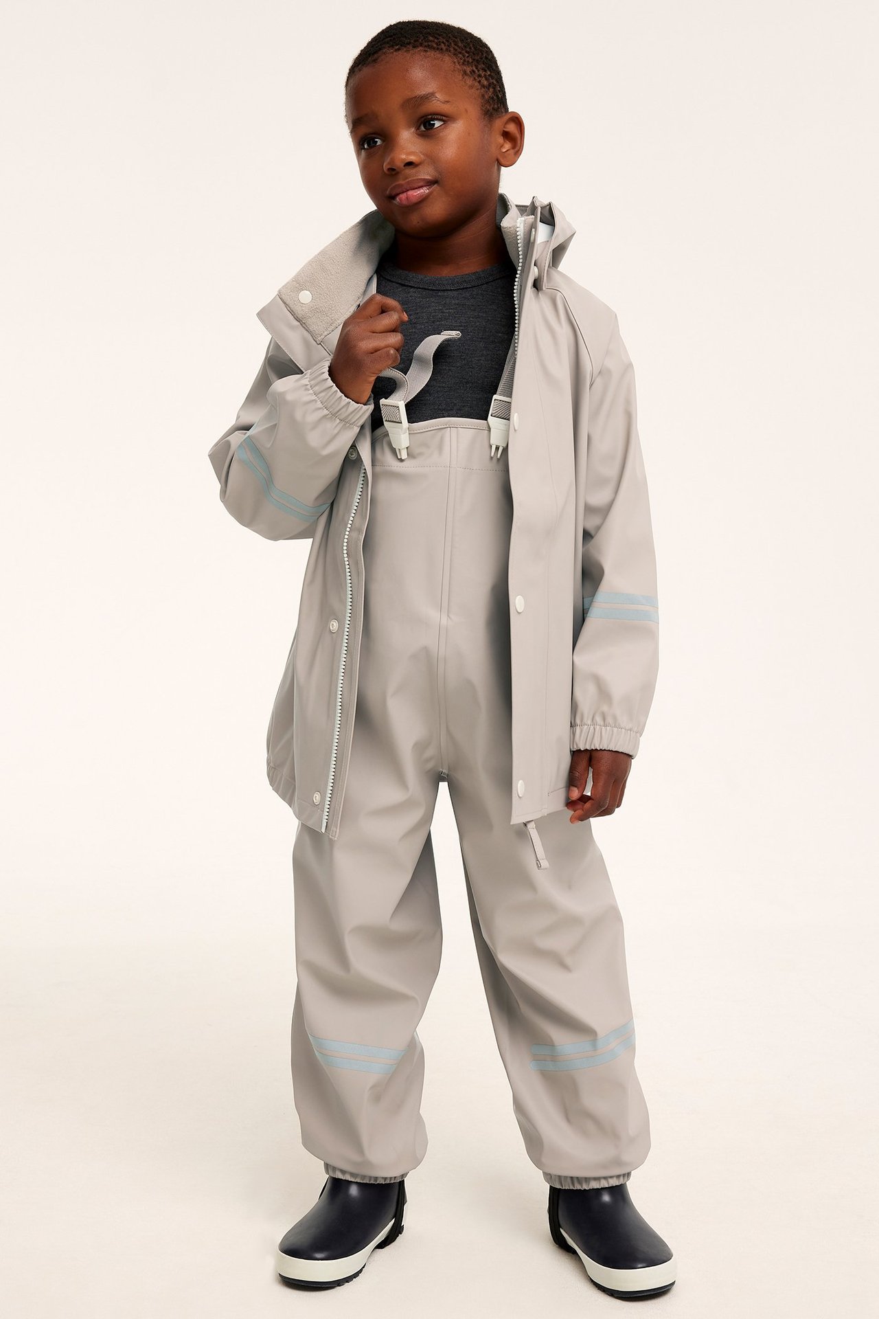 Barn bär beiga regnbyxor med hängslen, en matchande regnjacka och mörkblå gummistövlar.