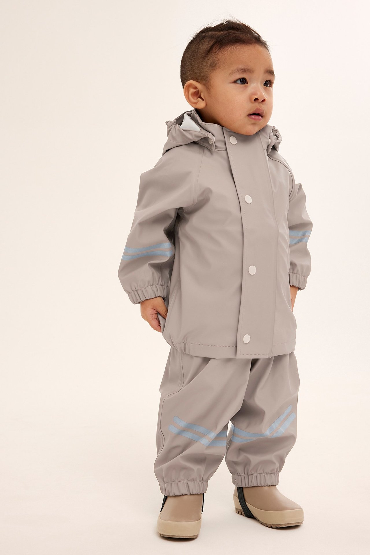 Framifrån, baby i beiga regnbyxor med reflexränder, matchande regnjacka och beiga regnstövlar.