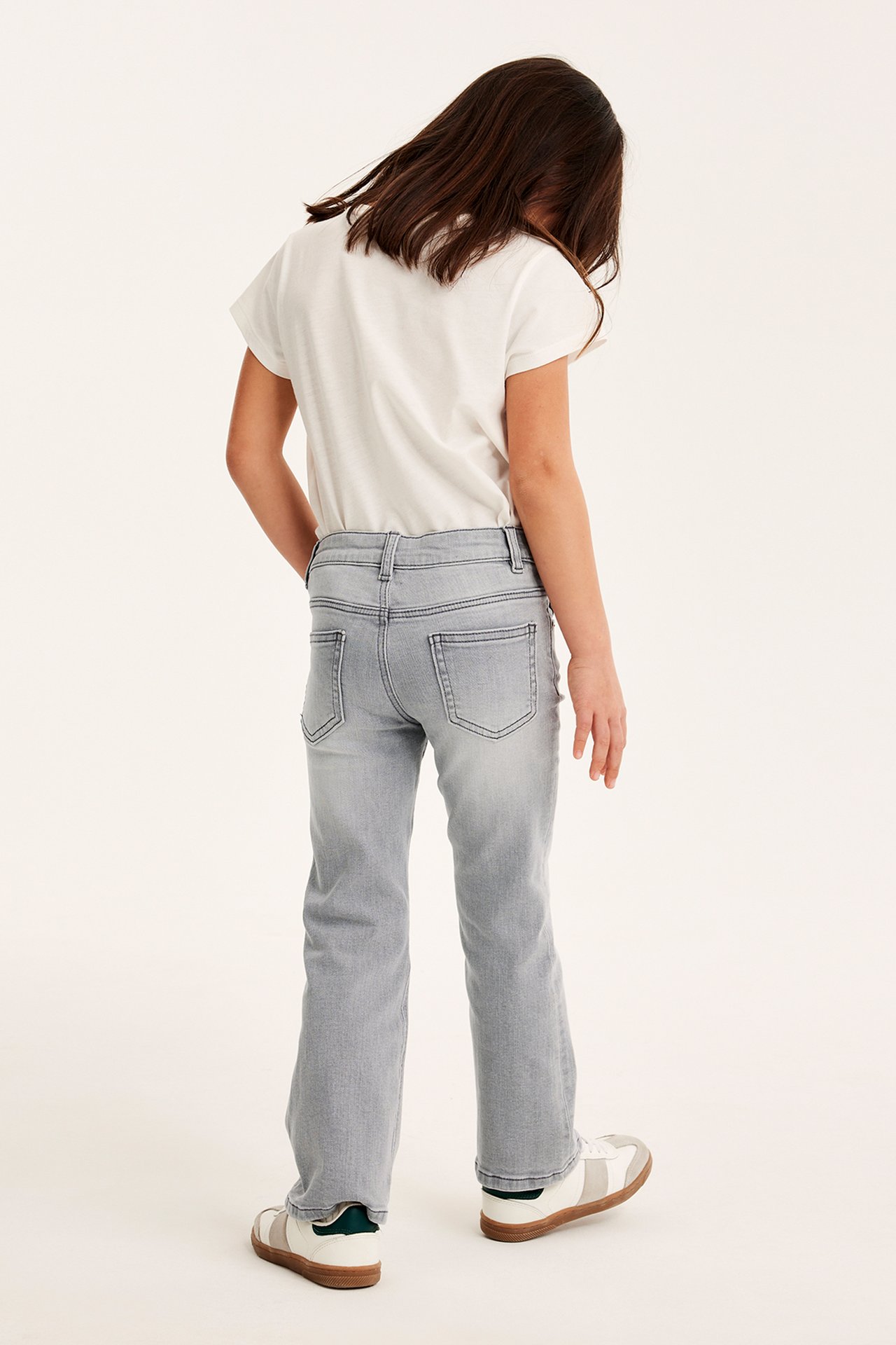 Bakifrån, ett barn i ljusgrå bootcut jeans i denim, en vit t-shirt och vita sneakers.